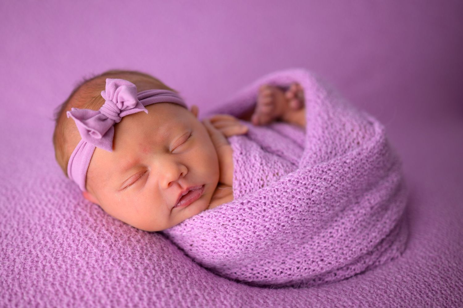 Newborn baby girl swaddled in purple knit wrap with bow headband, sleeping peacefully on lavender background.