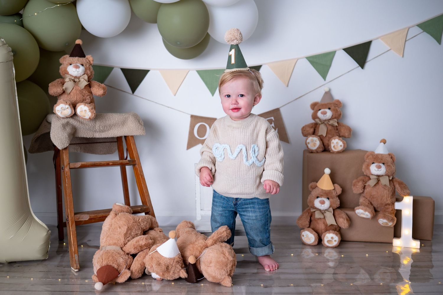 Toddler in party hat celebrates first birthday surrounded by teddy bears, balloons, and festive decorations.