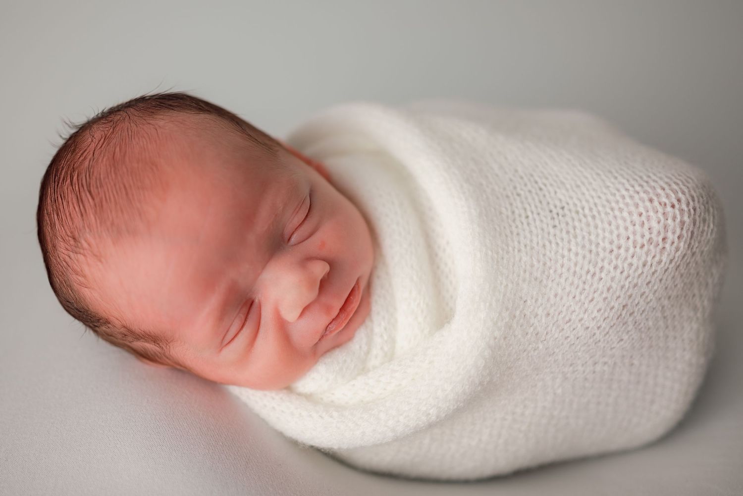 Peaceful newborn baby swaddled in a soft white knit wrap, sleeping soundly against a light gray background.