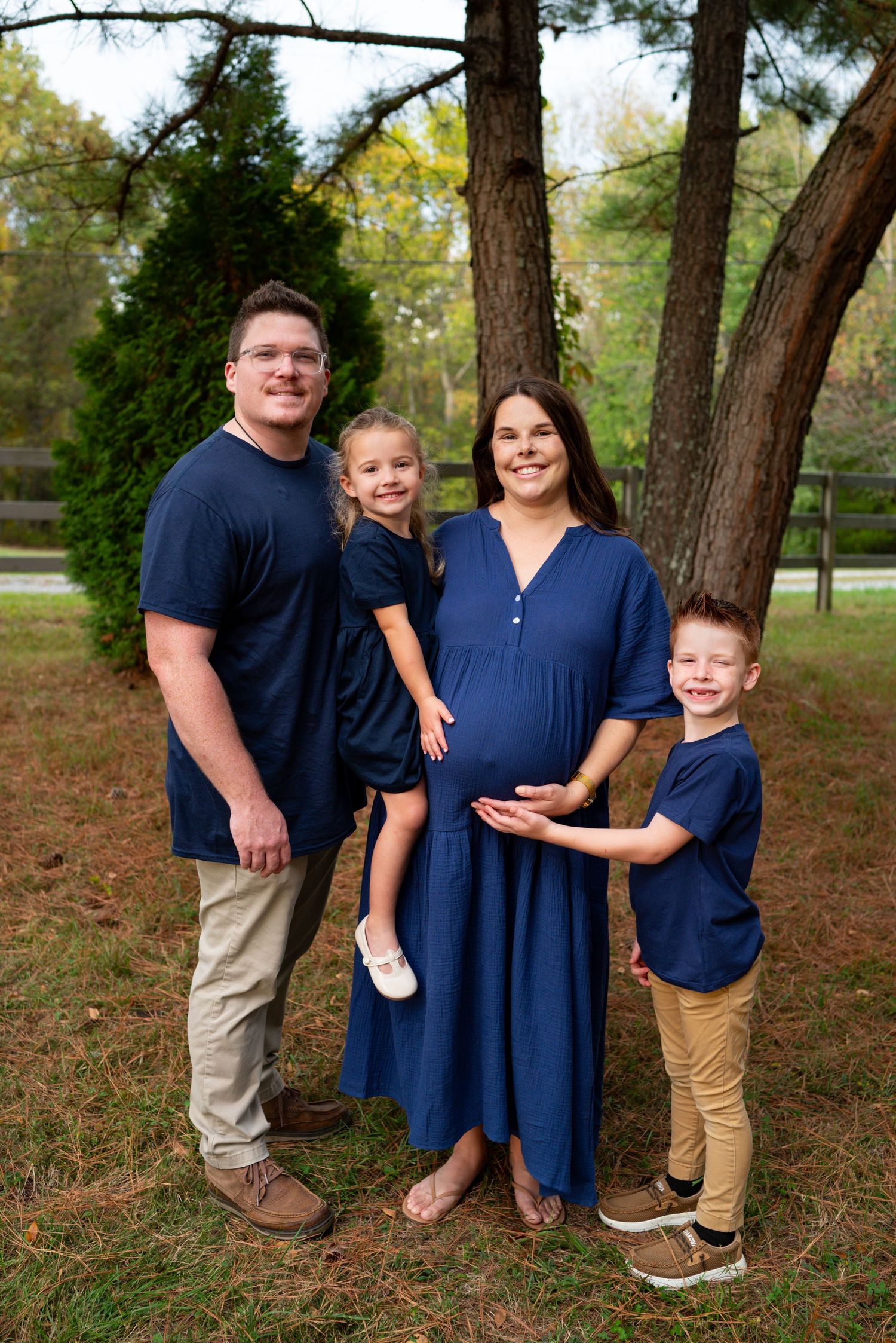 A happy family of four poses outdoors for maternity photos, all wearing coordinating navy and tan outfits.