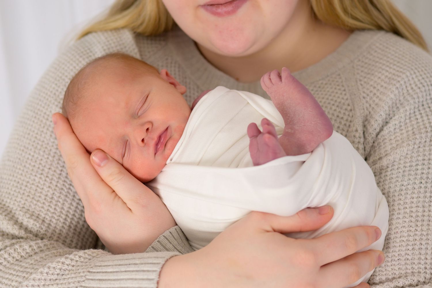 Mother in beige sweater gently holding swaddled sleeping newborn baby against her chest.
