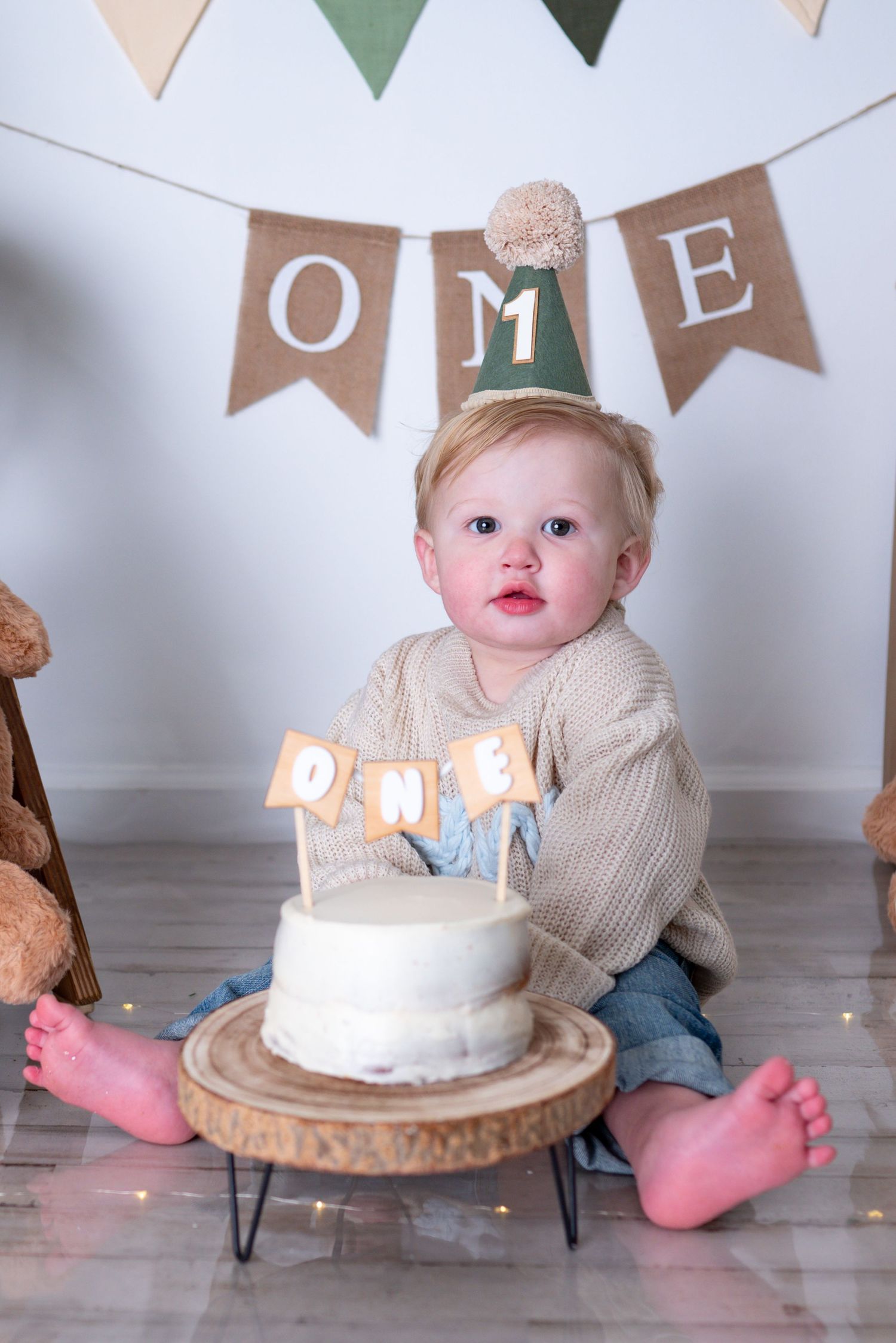 Baby boy in beige sweater sits beside first birthday smash cake with ONE topper and party hat.