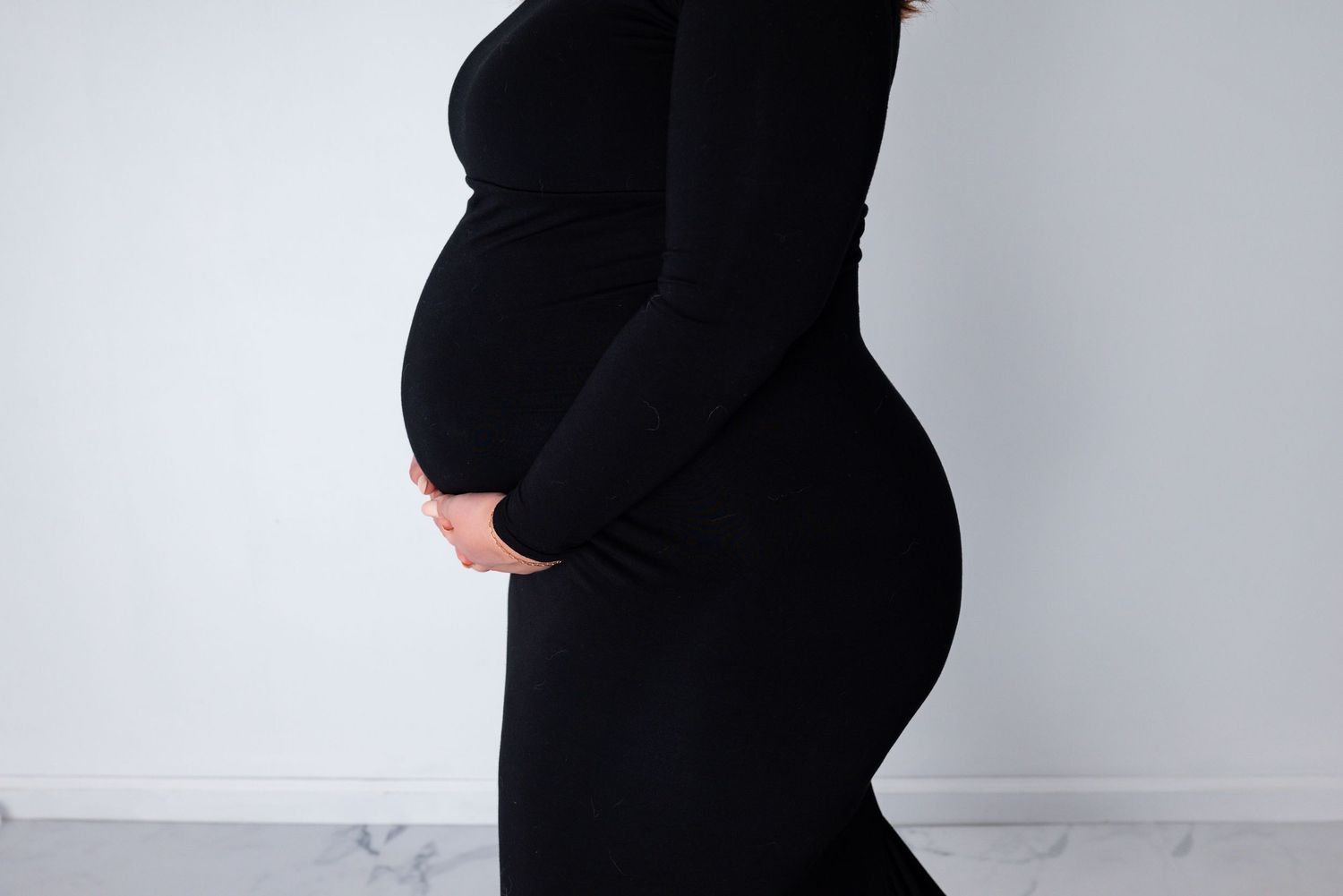 A side profile shot of a pregnant woman in a fitted black dress highlighting her baby bump in a white studio.