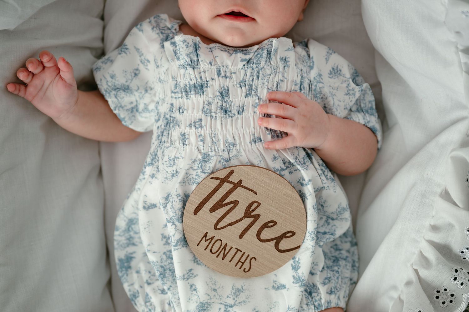 Baby lying on white bedding wearing floral outfit, holding a wooden three months milestone disc.