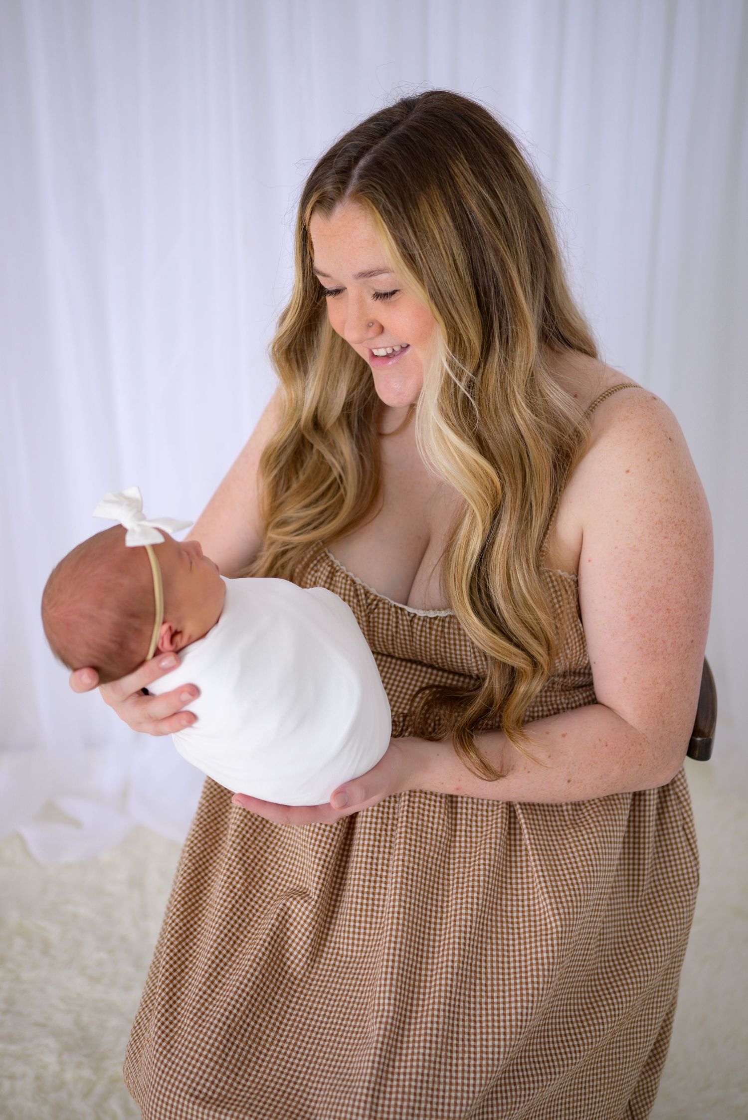 Smiling young mother in brown dress lovingly holding her swaddled newborn baby with white bow headband.