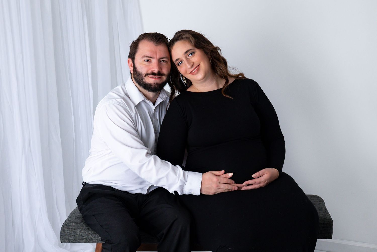 A couple poses together for maternity photos, with the pregnant woman in black and her partner in a white shirt.