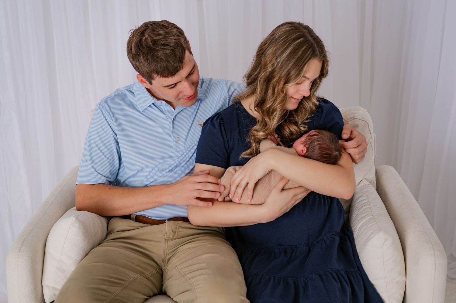 New parents sitting in white chair admiring their newborn baby during professional studio photography session.