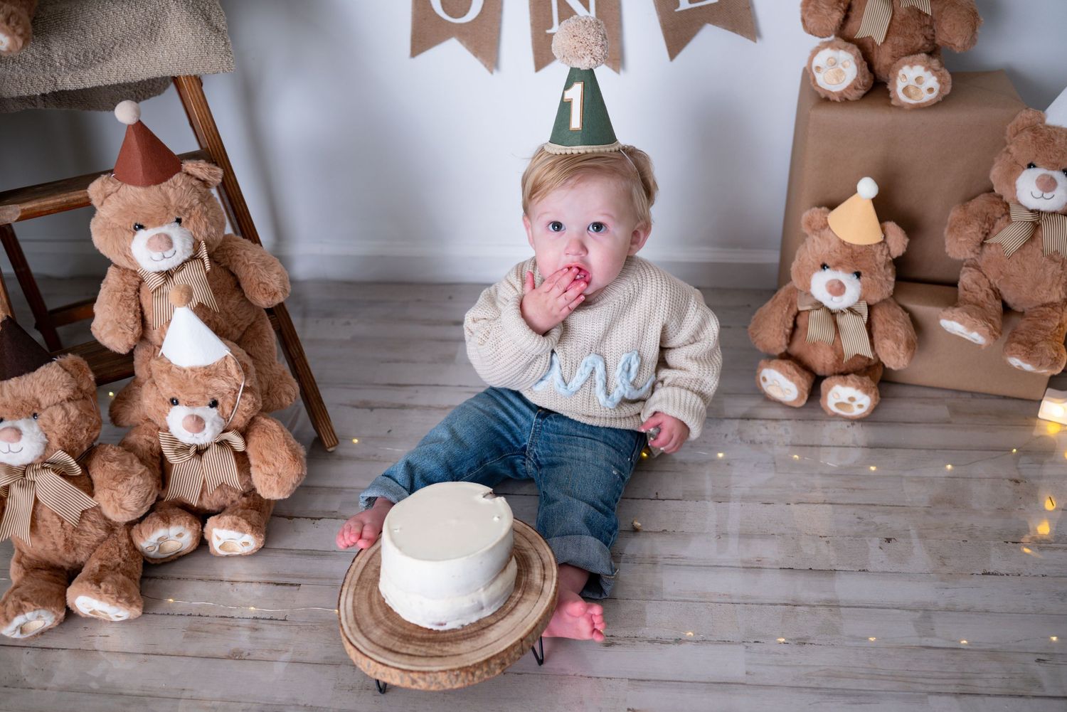 Baby boy in party hat eats cake with hands during first birthday smash cake session with teddy bears.
