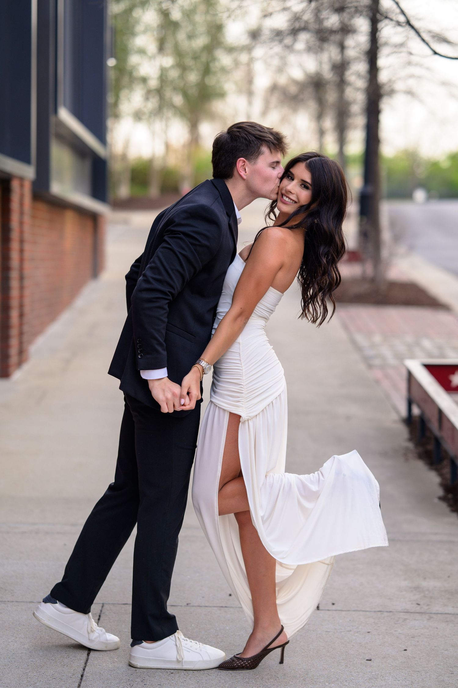 Man in black suit kissing smiling woman in white slit dress during outdoor engagement session.