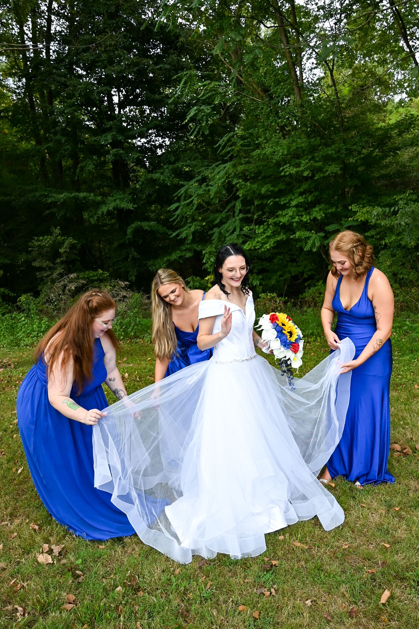 Bride in white gown holds colorful bouquet while three bridesmaids in royal blue dresses help arrange her train outdoors.