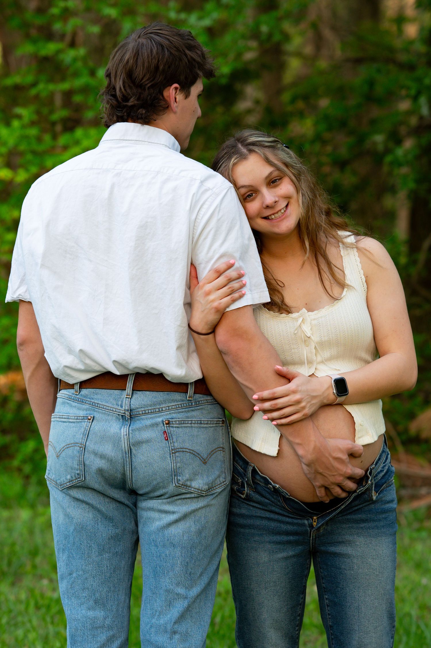 Pregnant woman smiling and embracing her partner outdoors, both wearing jeans, as he tenderly holds her baby bump.