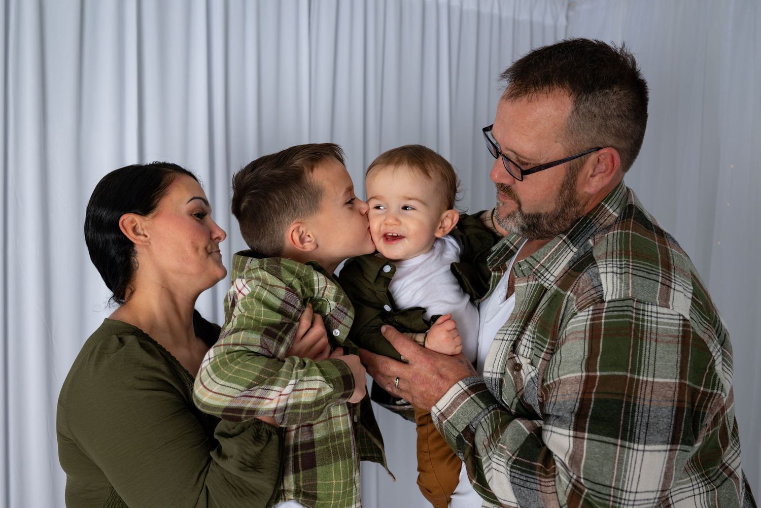 Happy family of four sharing a joyful moment as parents kiss older child while holding smiling baby indoors.