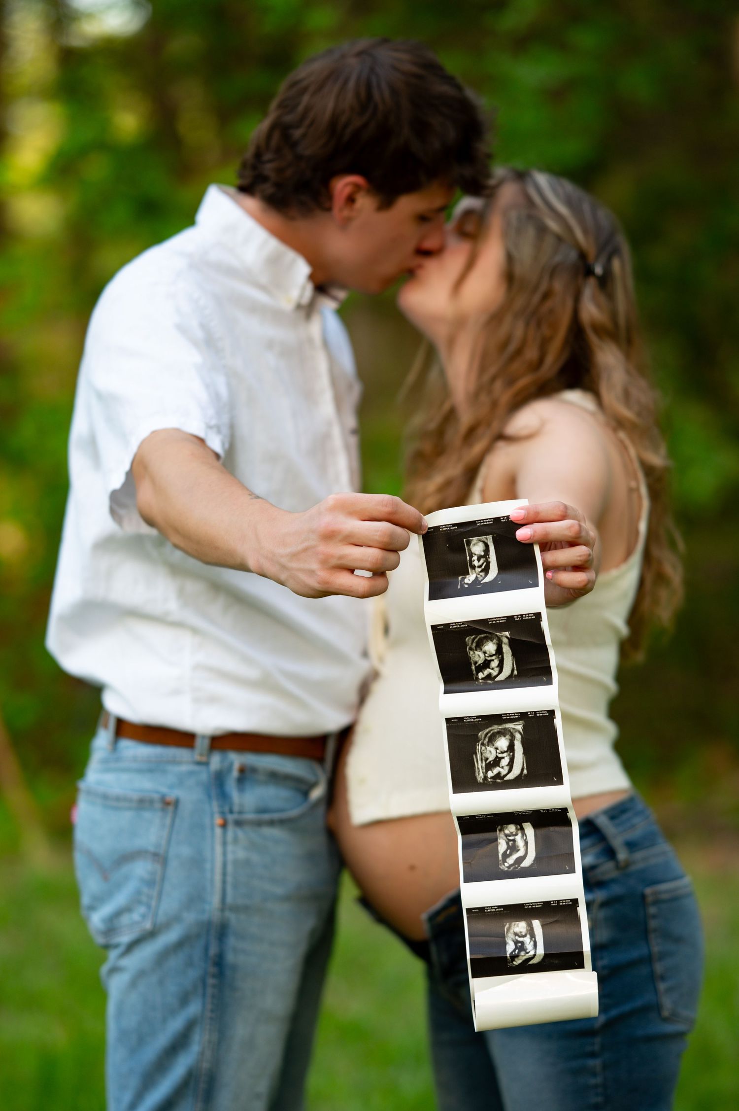 Expecting couple kissing while holding ultrasound photos during outdoor maternity photoshoot.