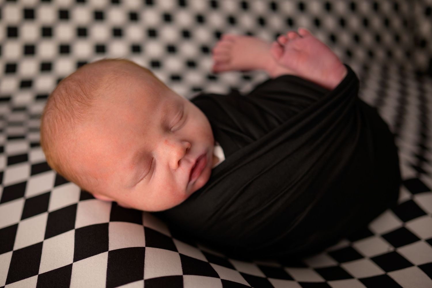Peaceful newborn baby swaddled in black wrap resting on checkered black and white blanket.
