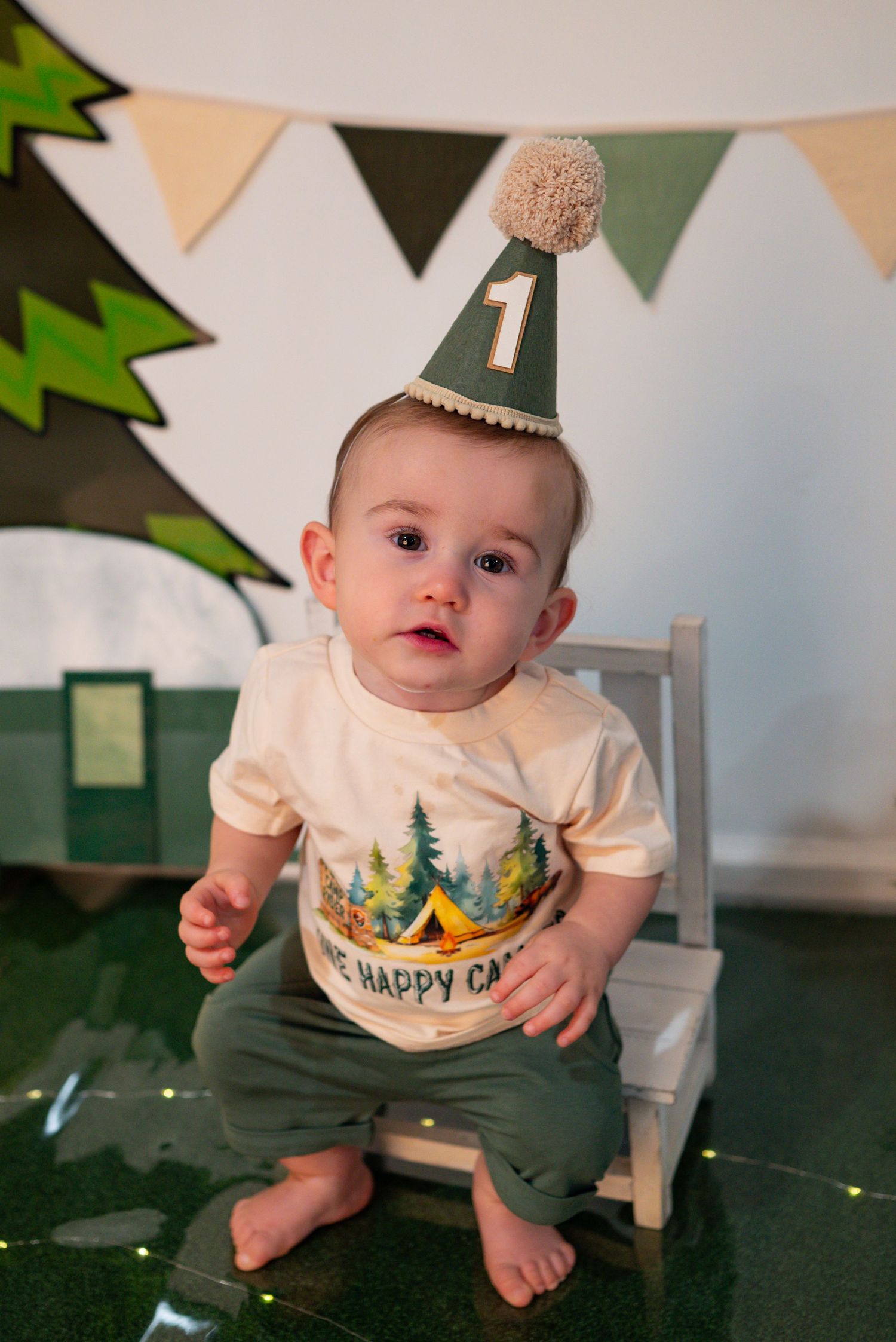 Baby boy in a camping-themed first birthday hat and One Happy Camper shirt sits for his 1st birthday photo.