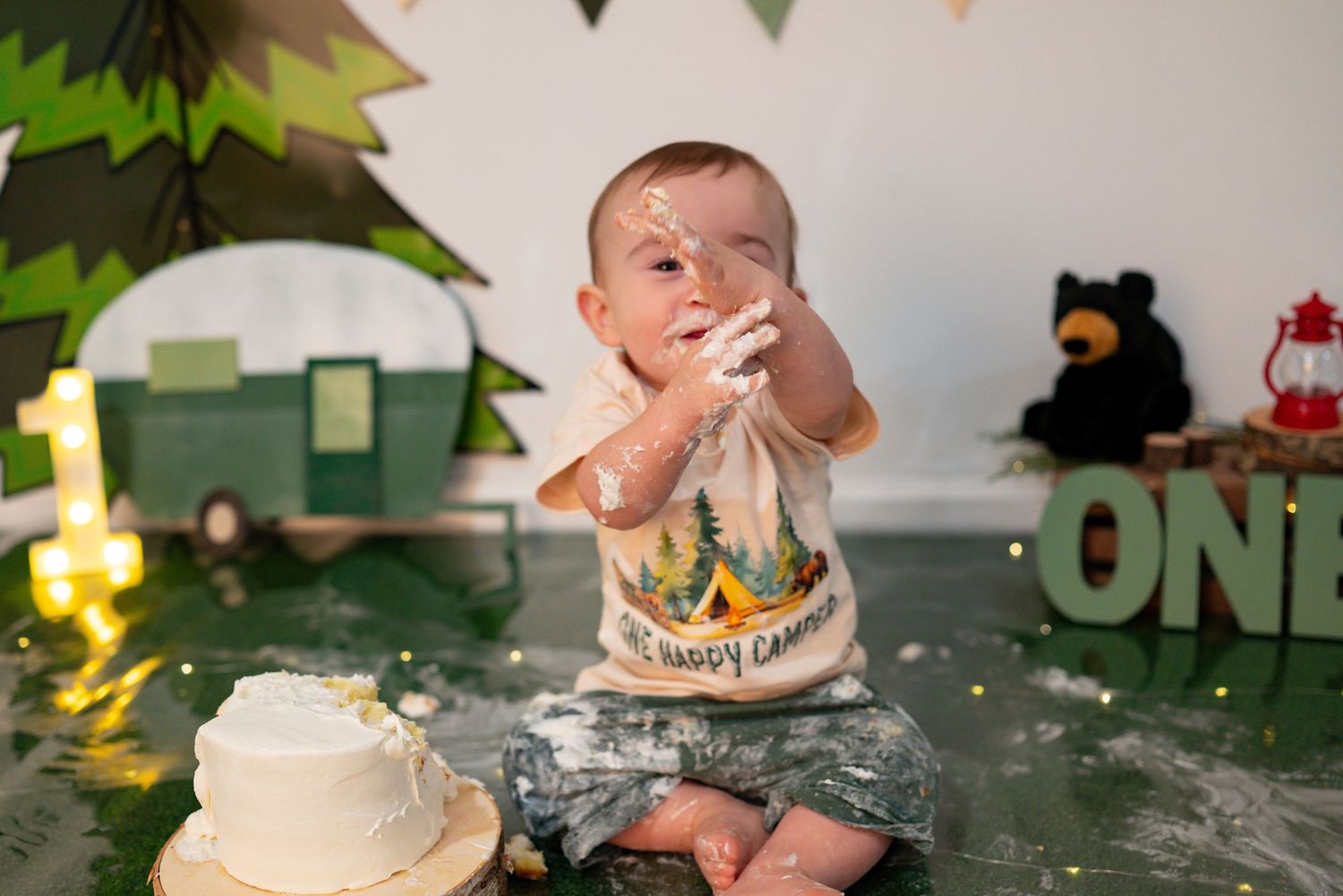 Baby boy holds frosting-covered hands up during his One Happy Camper first birthday cake smash photoshoot.
