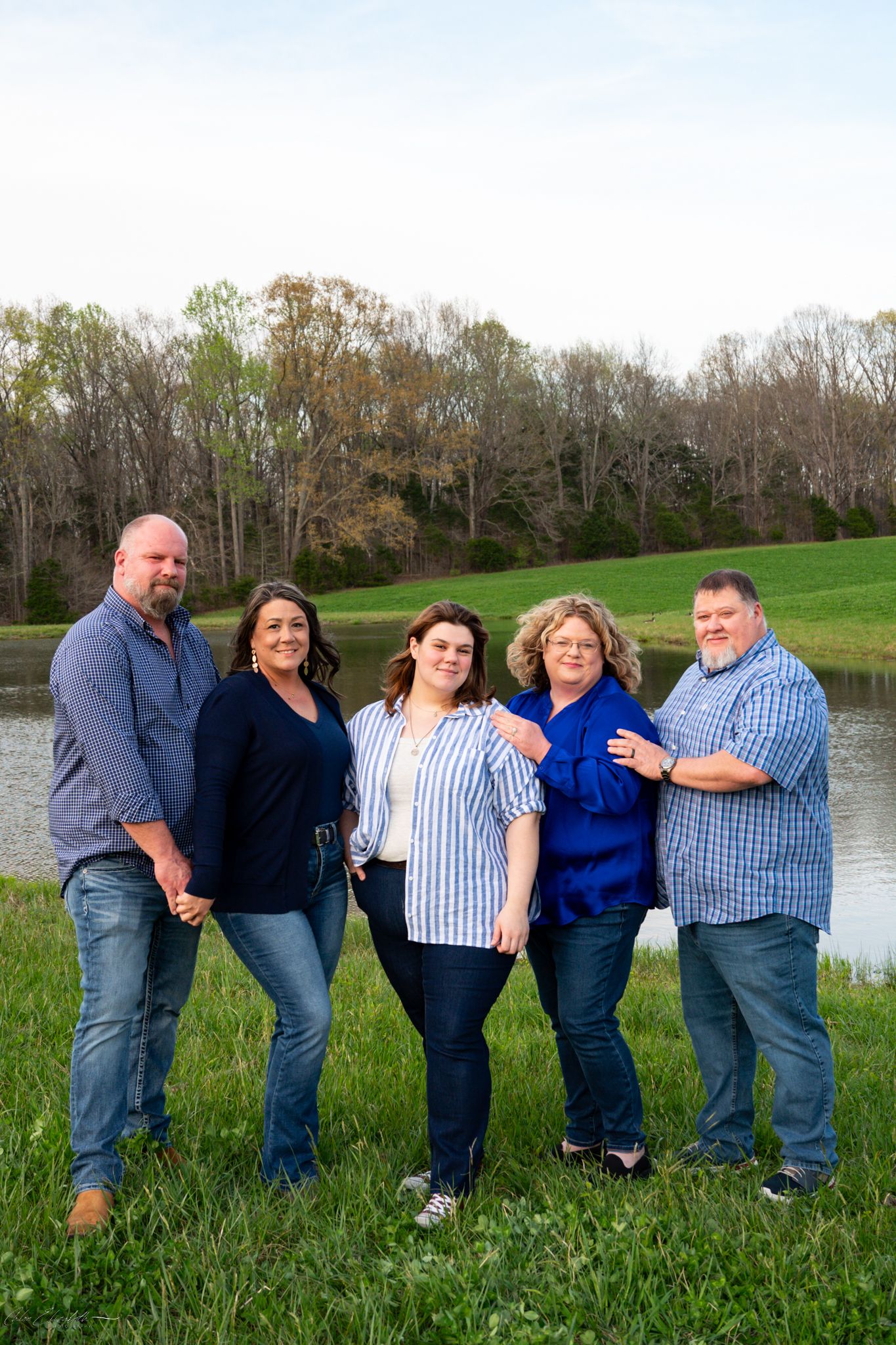 A group of five adults smiles together for a family portrait beside a peaceful pond surrounded by spring trees.