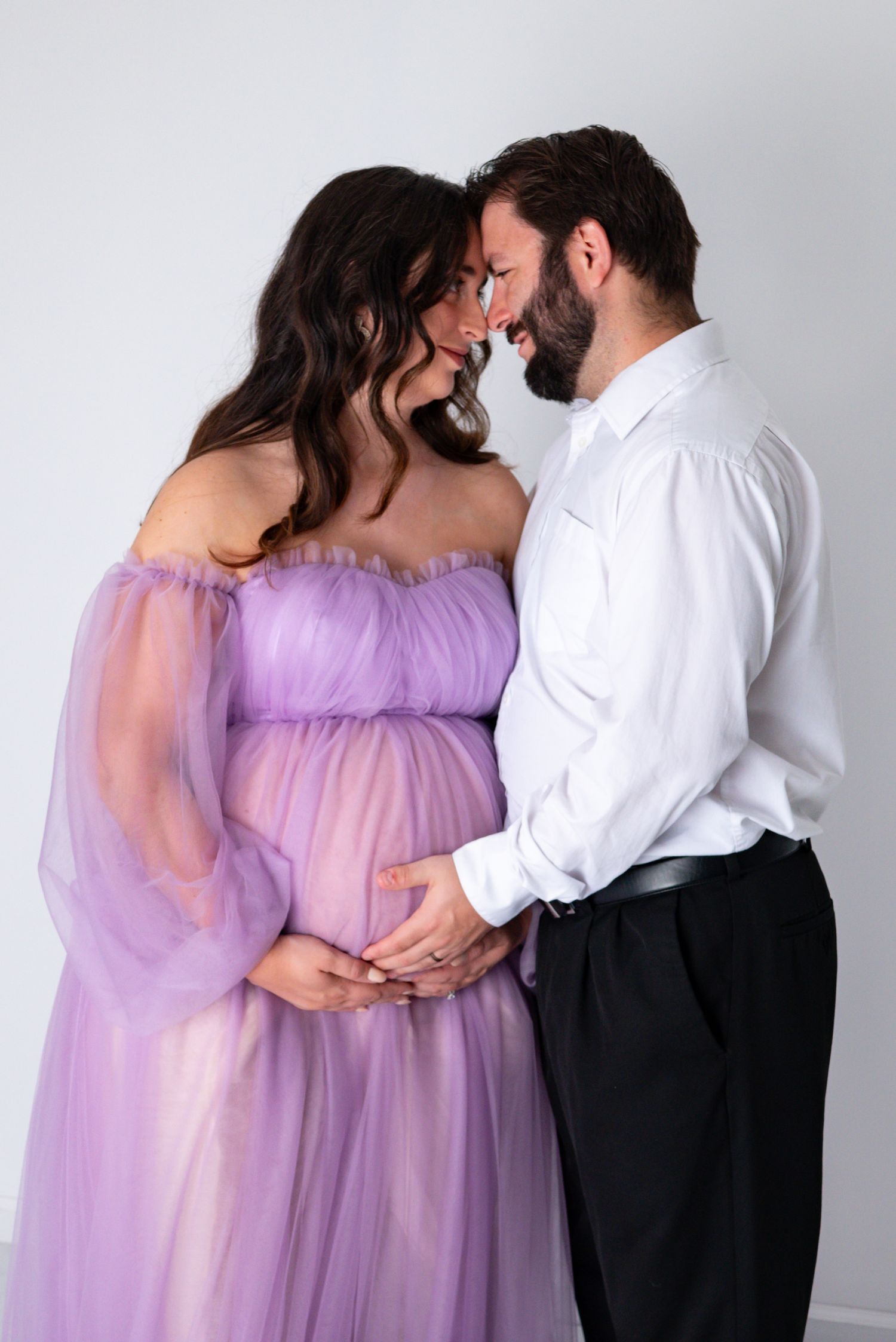 A couple shares an intimate forehead-to-forehead moment during a studio maternity session in elegant attire.
