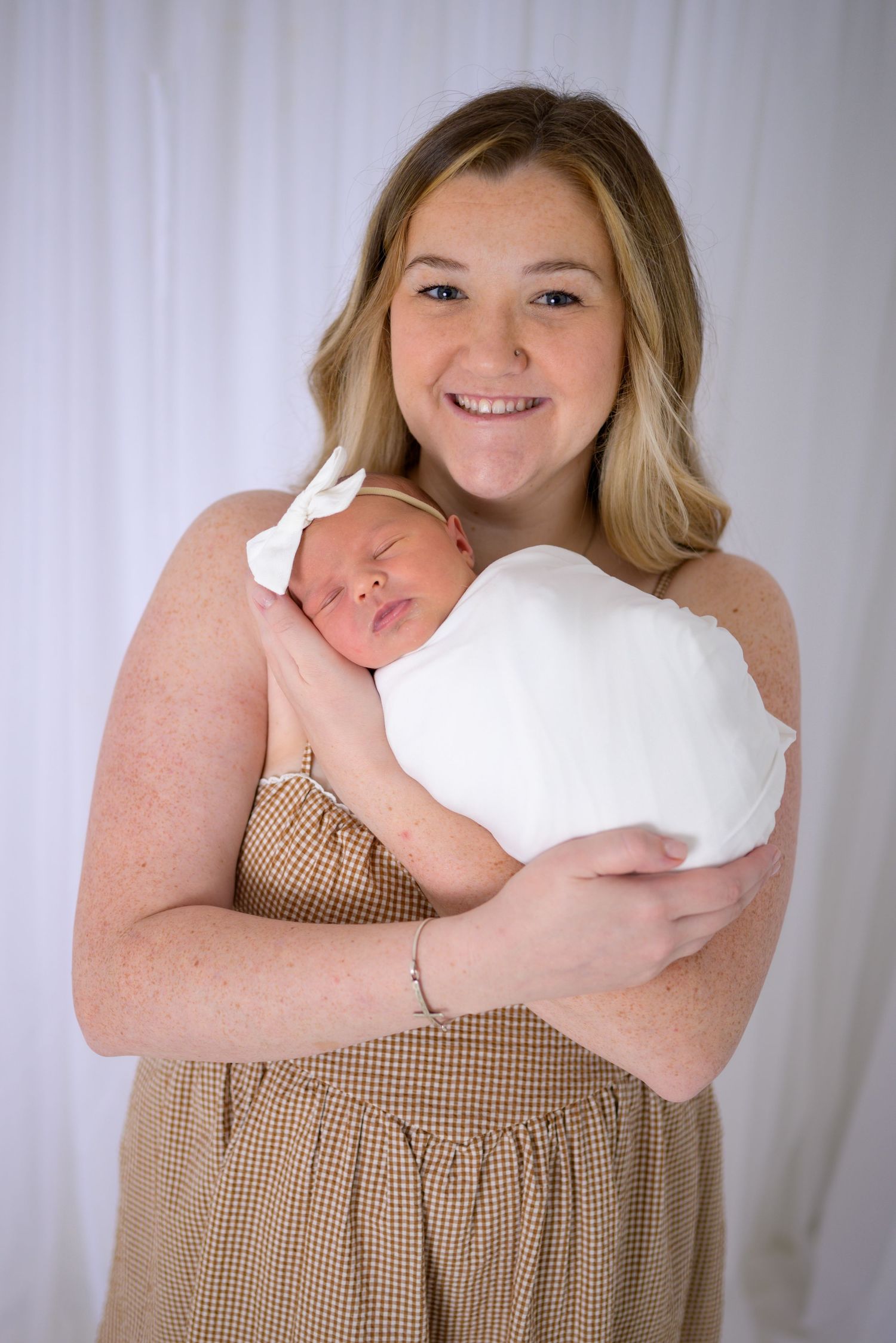 Smiling young mother in brown dress proudly holding her sleeping newborn wrapped in white swaddle with bow.