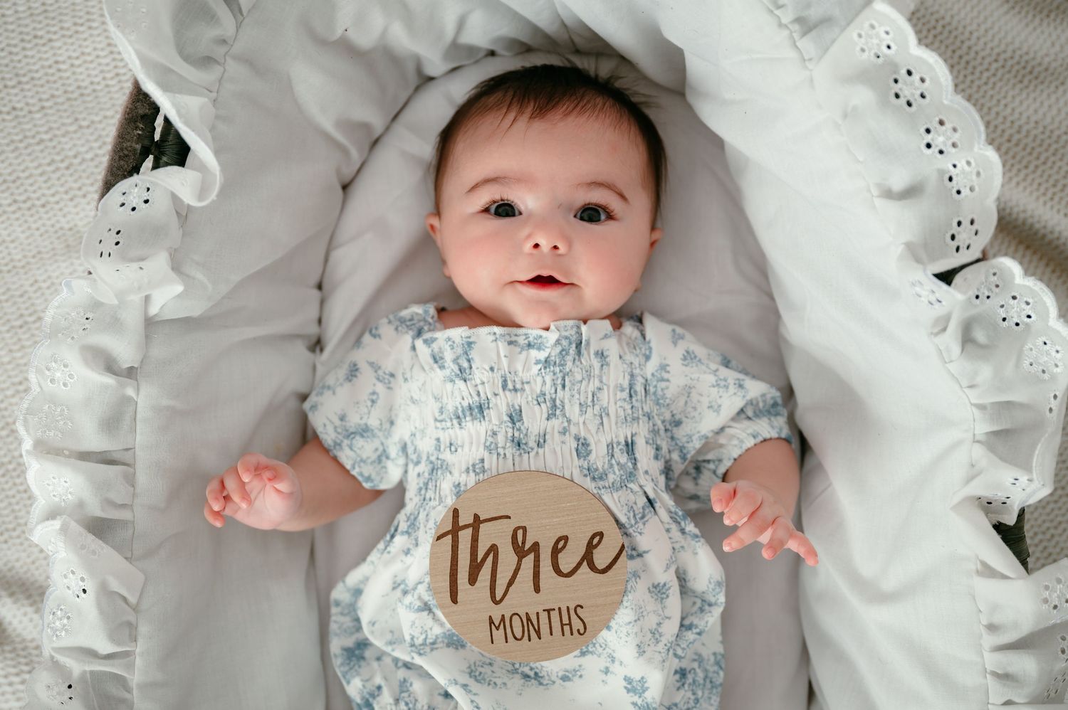 A three-month-old baby lying in a white bassinet wearing a floral onesie with a wooden milestone disc.