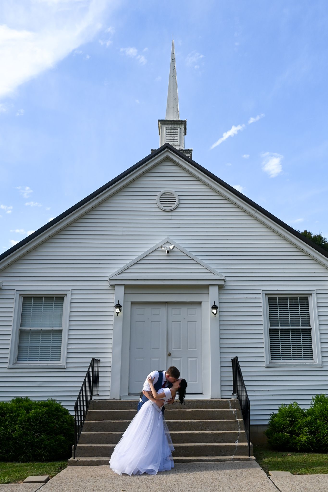 Bride and groom share a romantic dip kiss on the steps of a white chapel with a steeple on their wedding day.