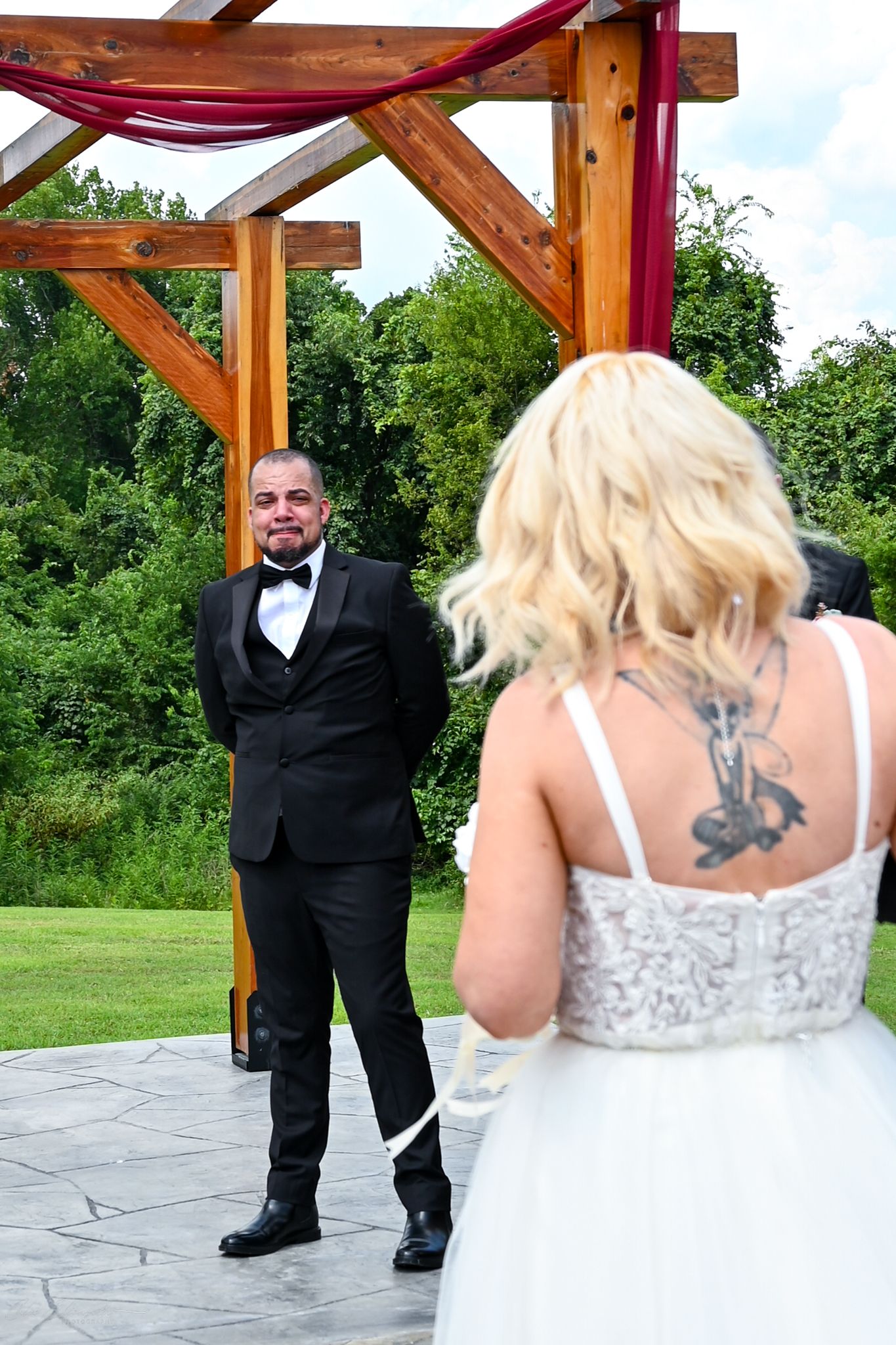 Groom in black tuxedo smiles at tattooed bride in white lace gown during outdoor wedding ceremony.