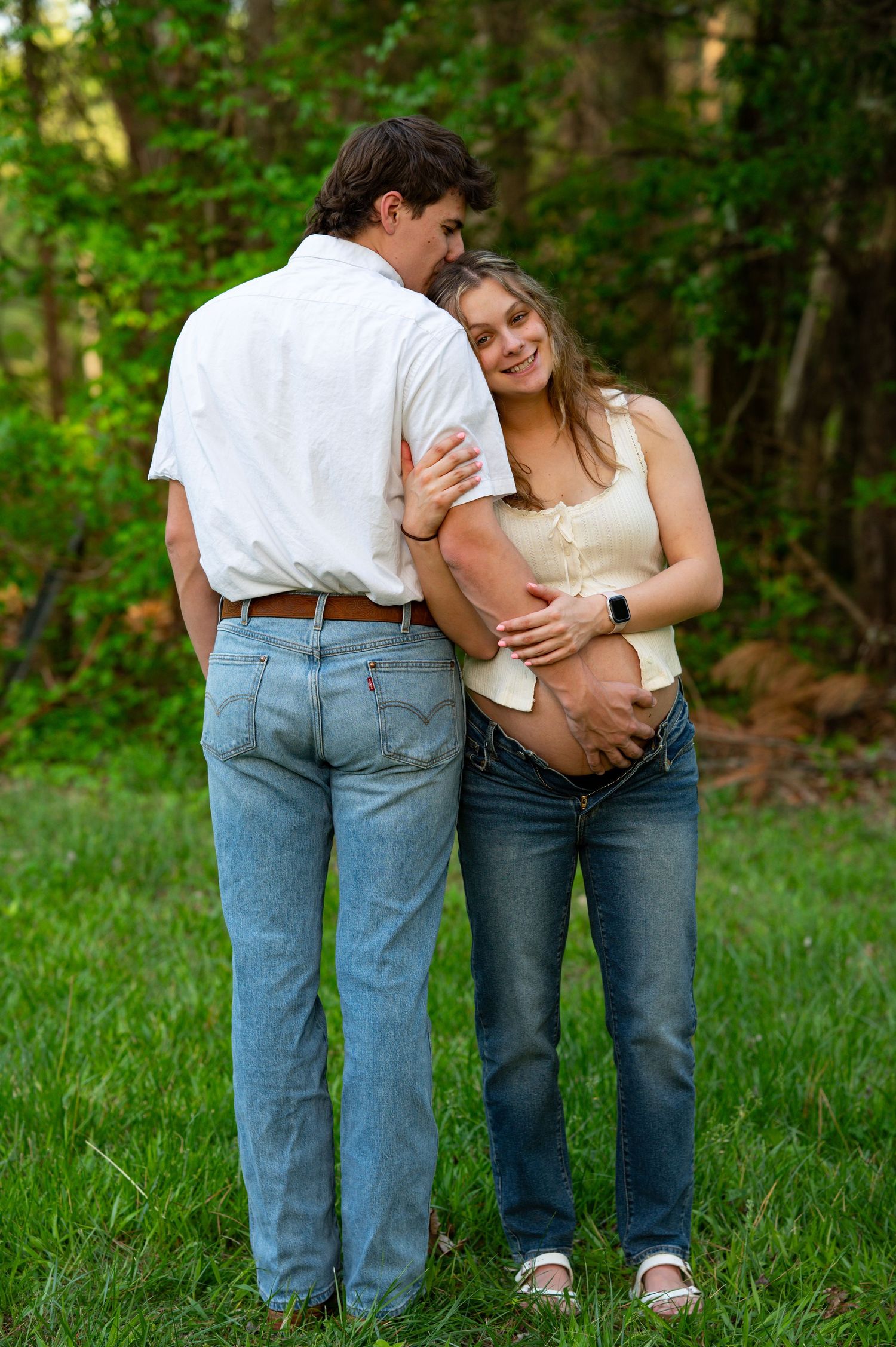Pregnant couple embracing outdoors in green field, man in white shirt and jeans, woman in beige top smiling warmly.