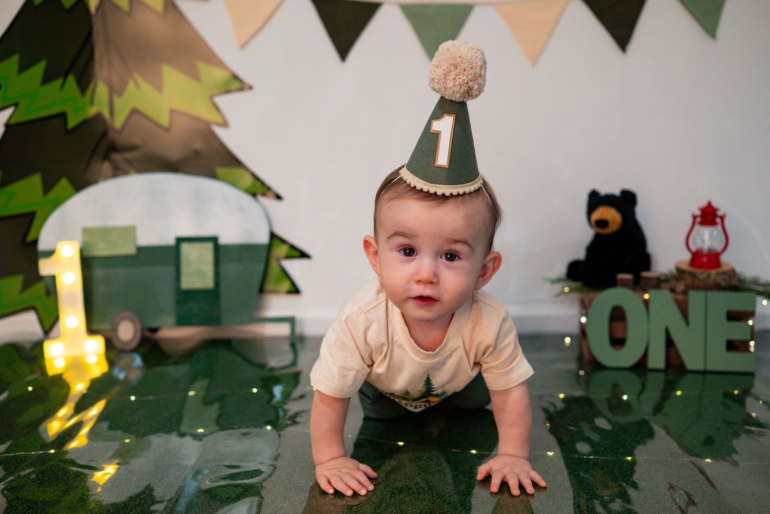 One-year-old baby crawls toward camera wearing a birthday hat at a camping-themed first birthday photoshoot.