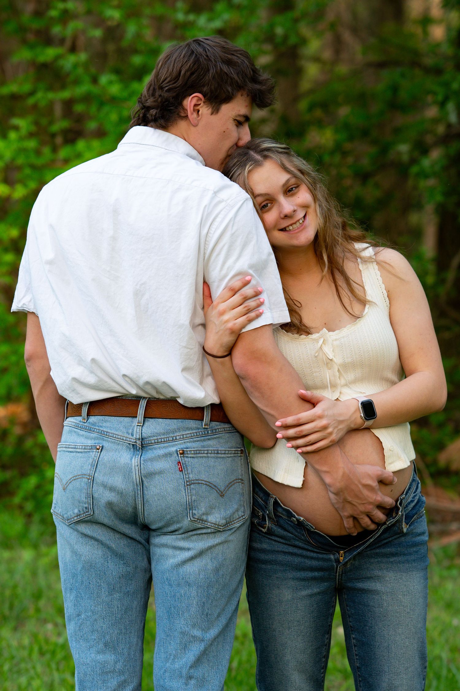 Happy pregnant woman leaning into her partner outdoors while he kisses her cheek and cradles her baby bump.