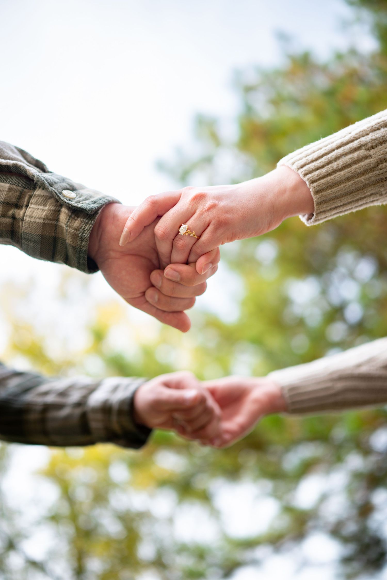 Close-up of a couple holding hands outdoors with green trees in the background during fall season.
