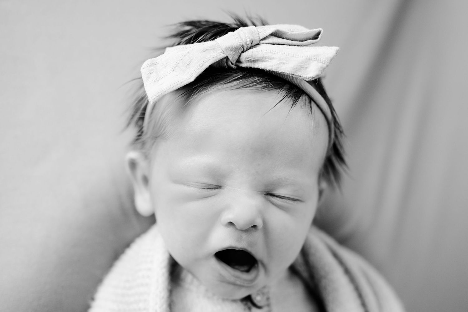 Black and white photo of yawning newborn baby wearing a bow headband.