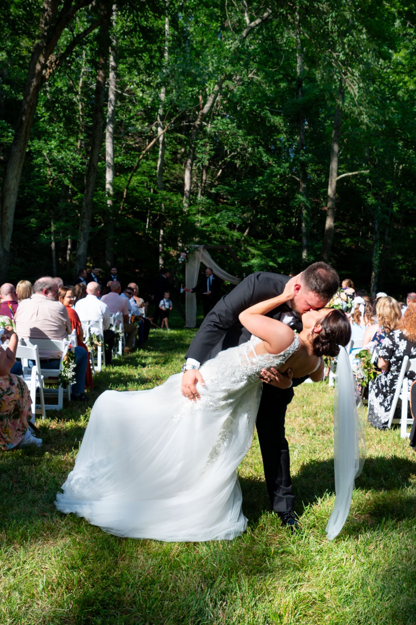 Groom dips bride for a kiss at outdoor wedding ceremony surrounded by guests and lush green trees.