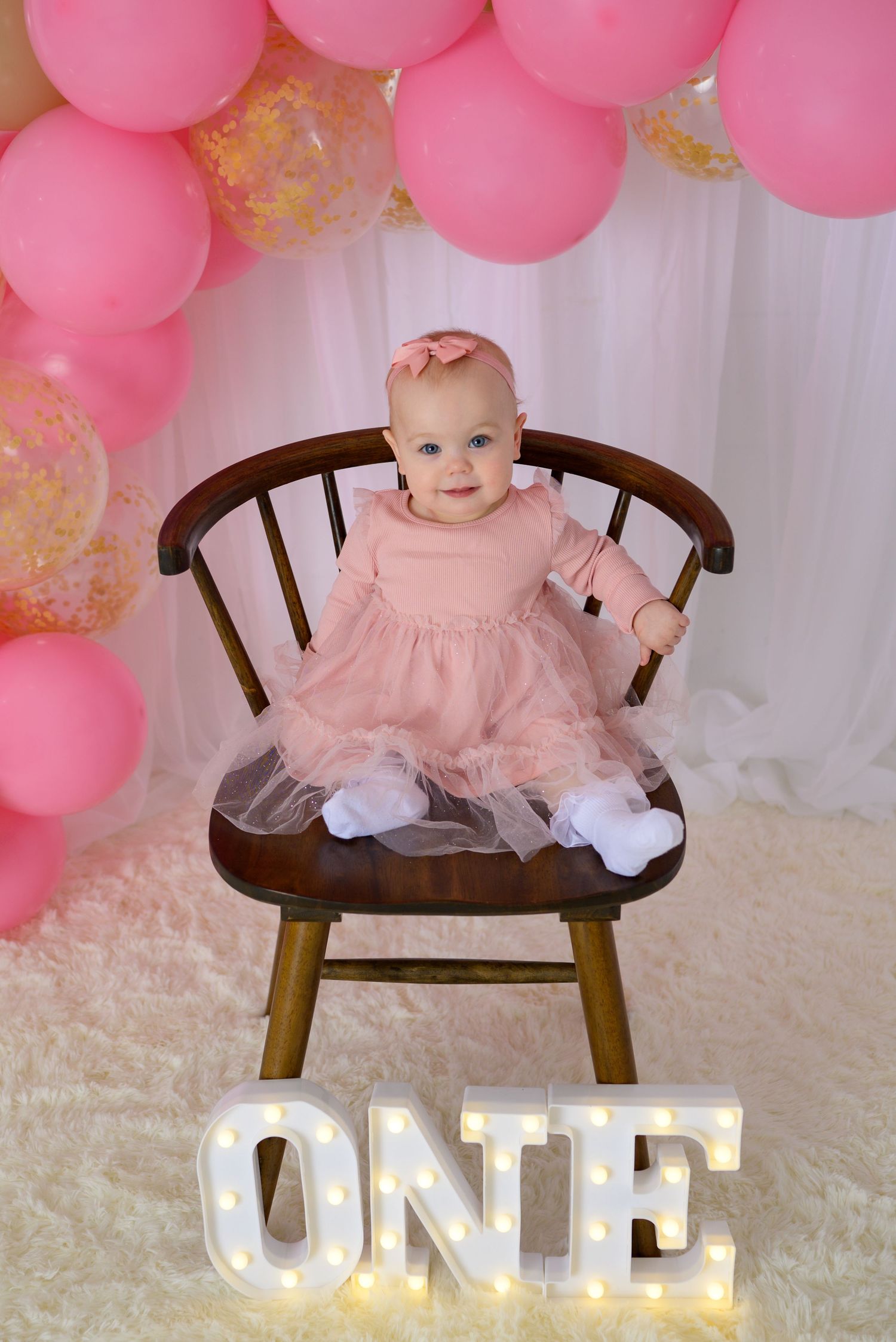 Baby girl in pink tulle dress on wooden chair with illuminated ONE sign and pink balloons.