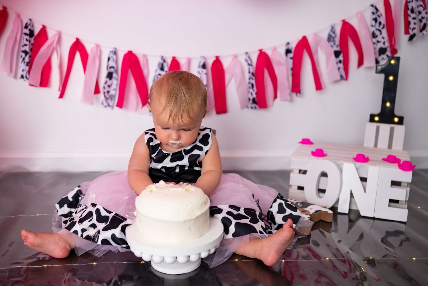 One-year-old girl in cow print dress explores white birthday cake at cake smash session.