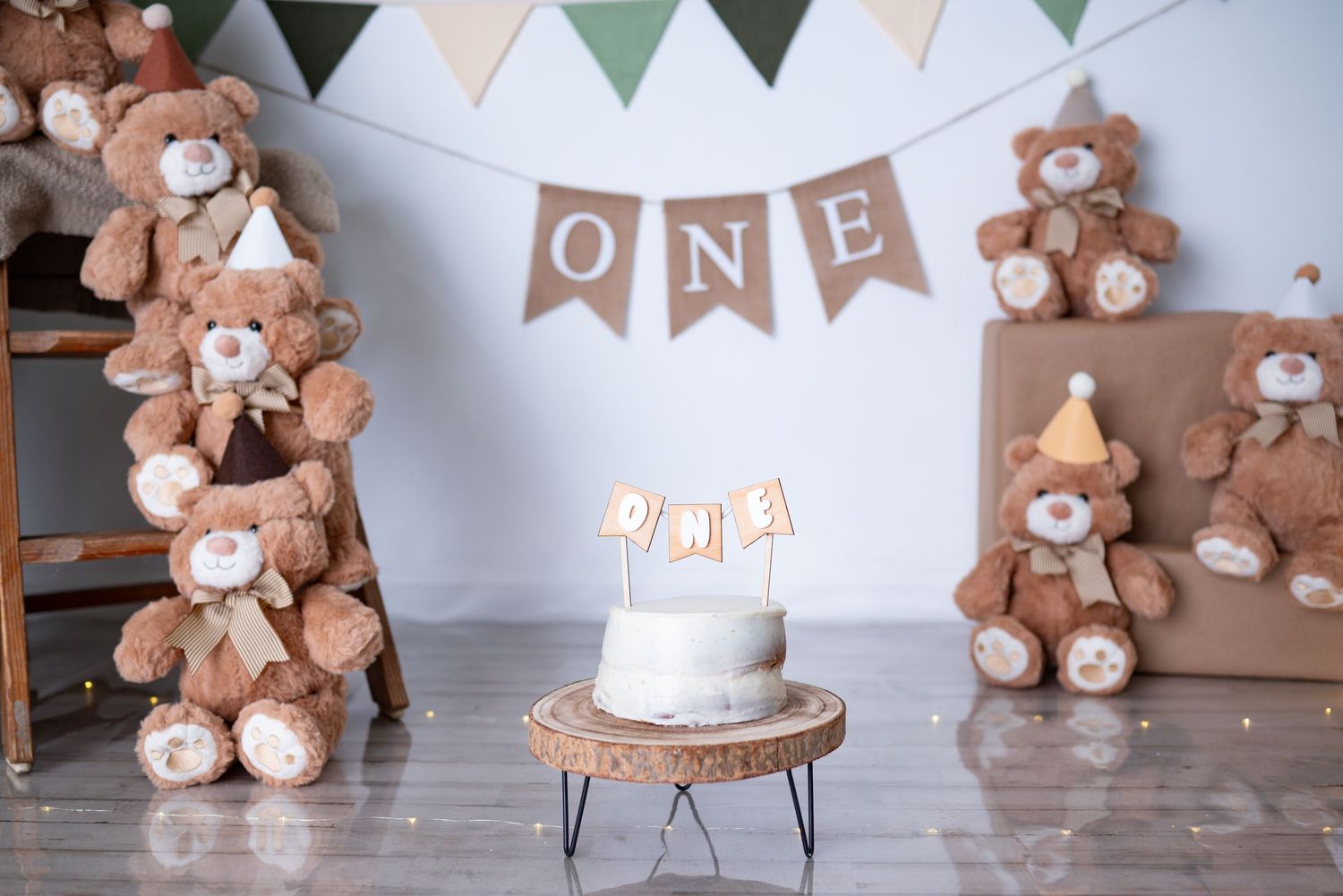 First birthday cake smash setup with teddy bears, ONE banner, and white cake on wood stand.