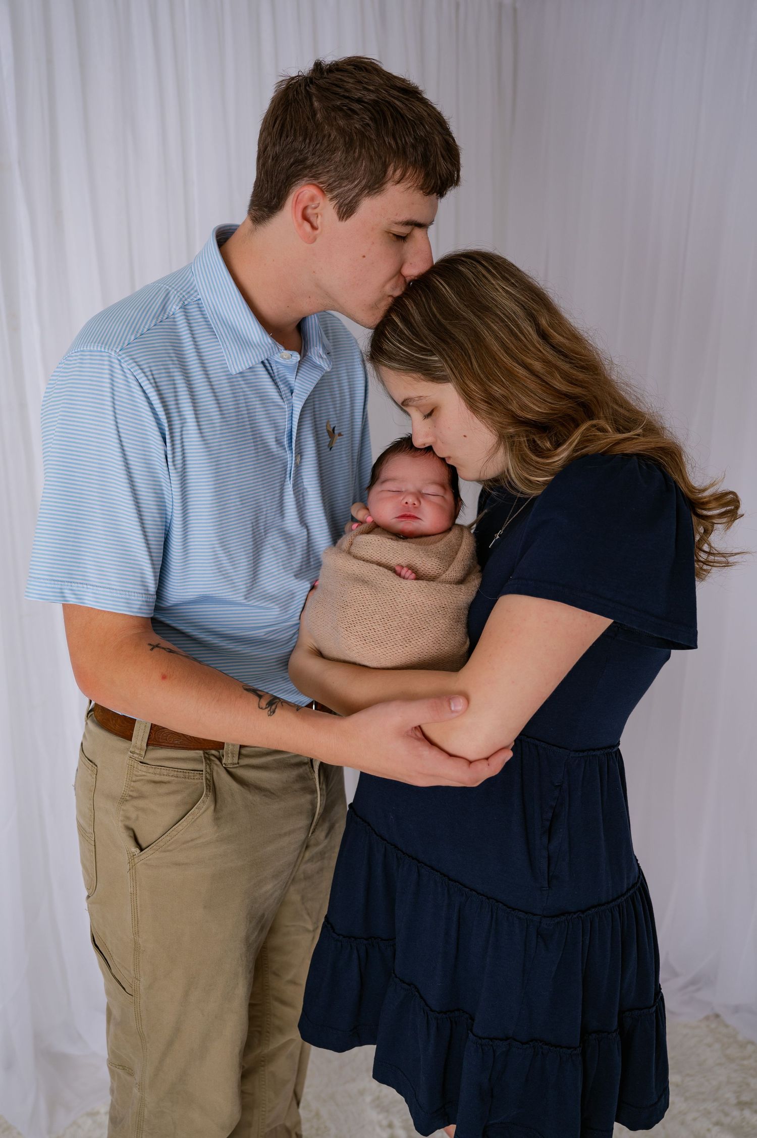 New parents share a tender moment kissing their newborn baby wrapped in a tan swaddle blanket.