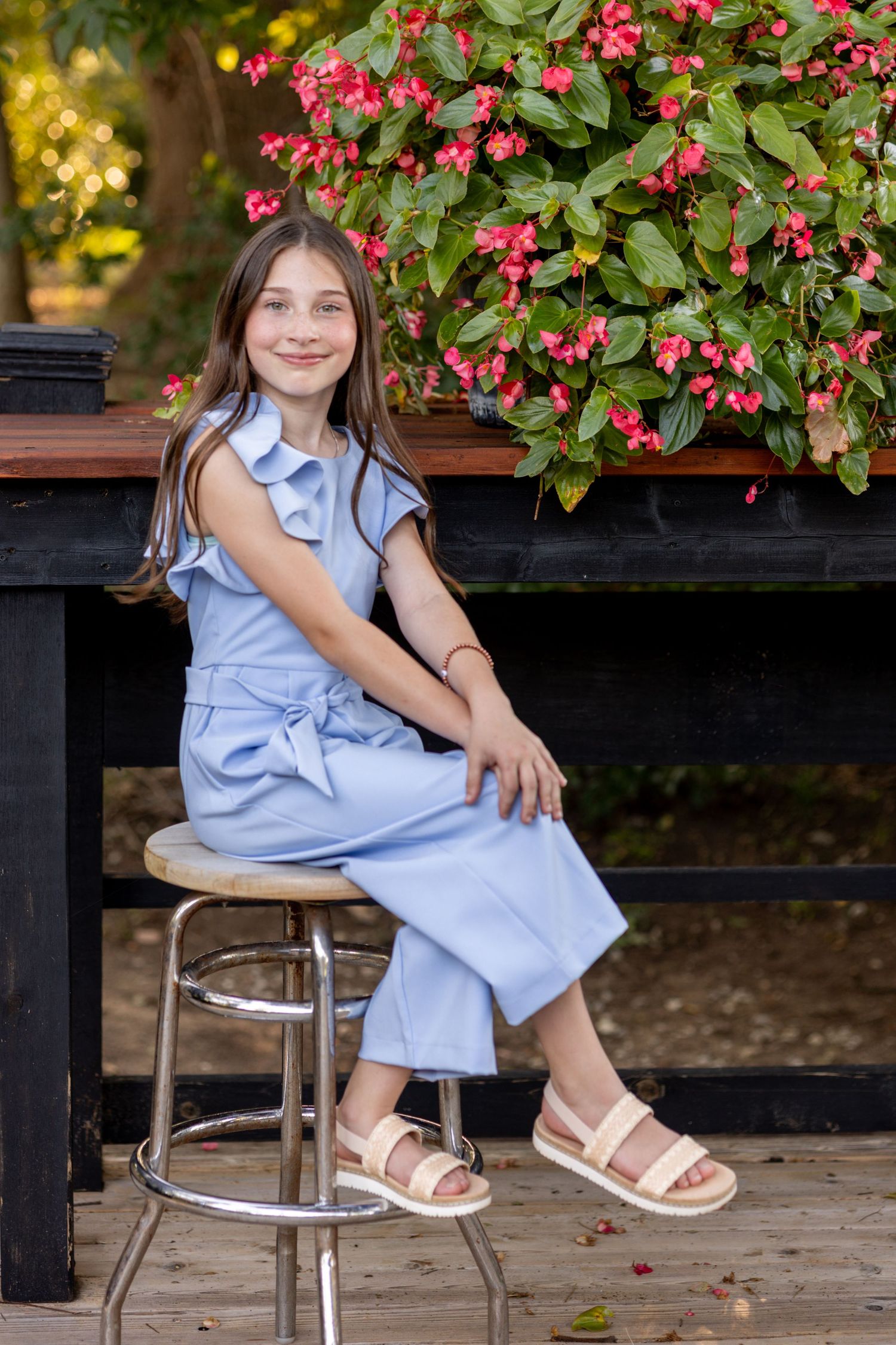 Young girl in blue jumpsuit sitting on a stool outdoors with pink flowers in background.