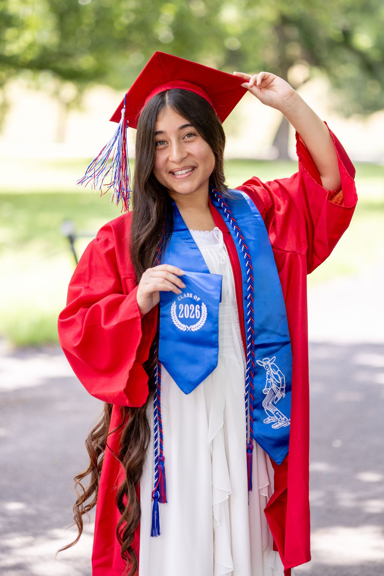 Young woman in red graduation cap and gown with blue stole reading Class of 2026, smiling outdoors.