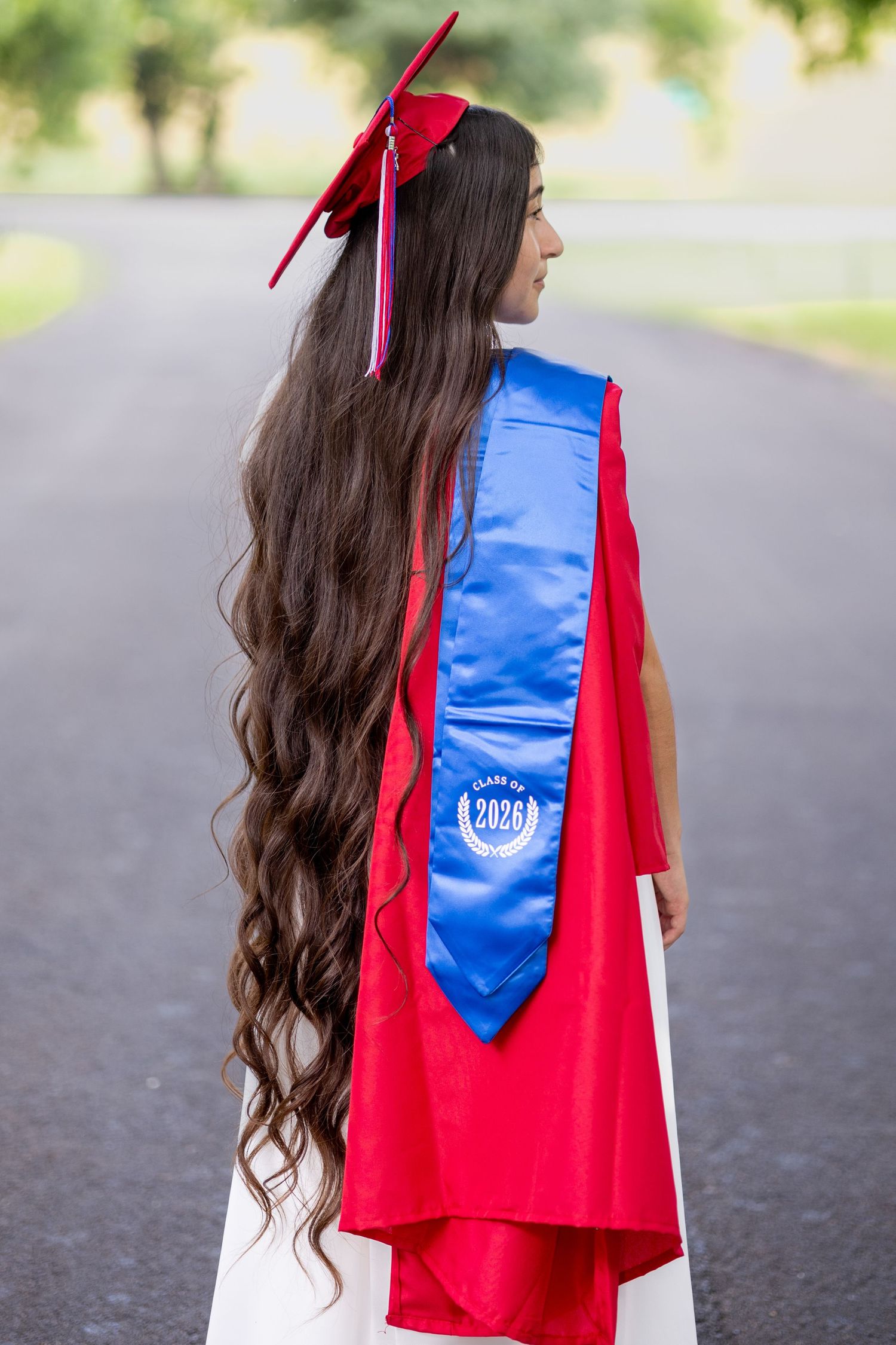 A young woman in red graduation cap and gown with blue Class of 2026 sash, showing long wavy hair, standing on a road.