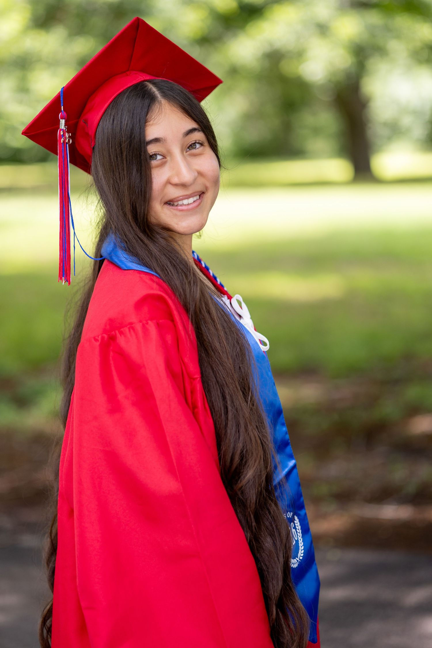 Smiling young woman in red graduation cap and gown poses outdoors in a lush green park setting.