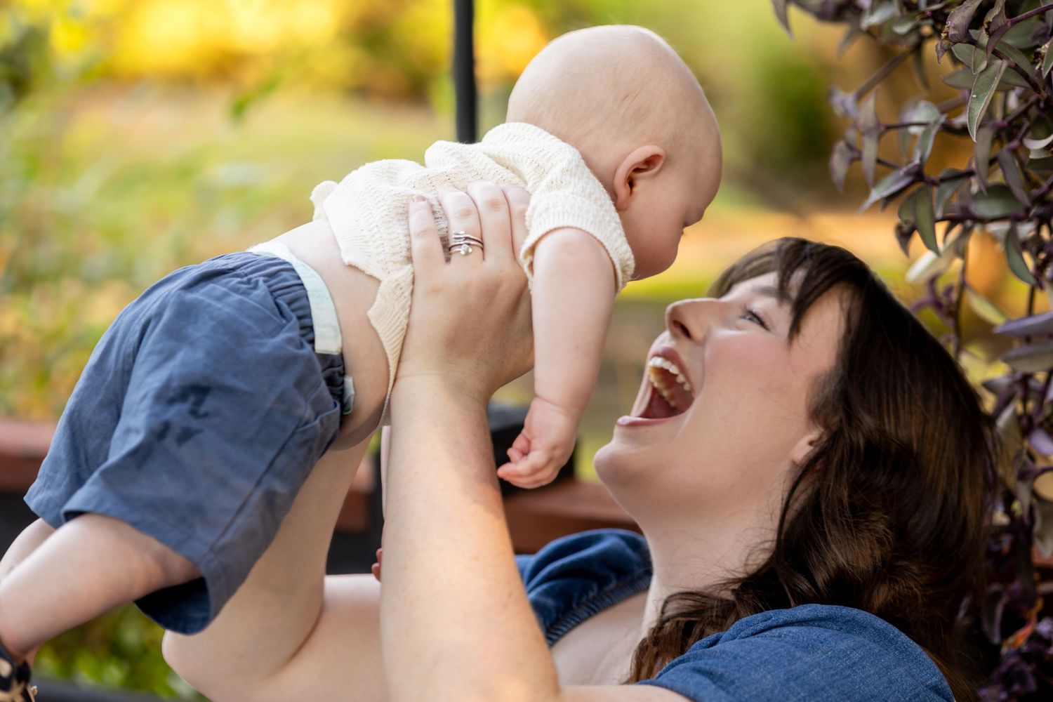 A laughing mother joyfully lifts her smiling baby in the air during an outdoor photo session.