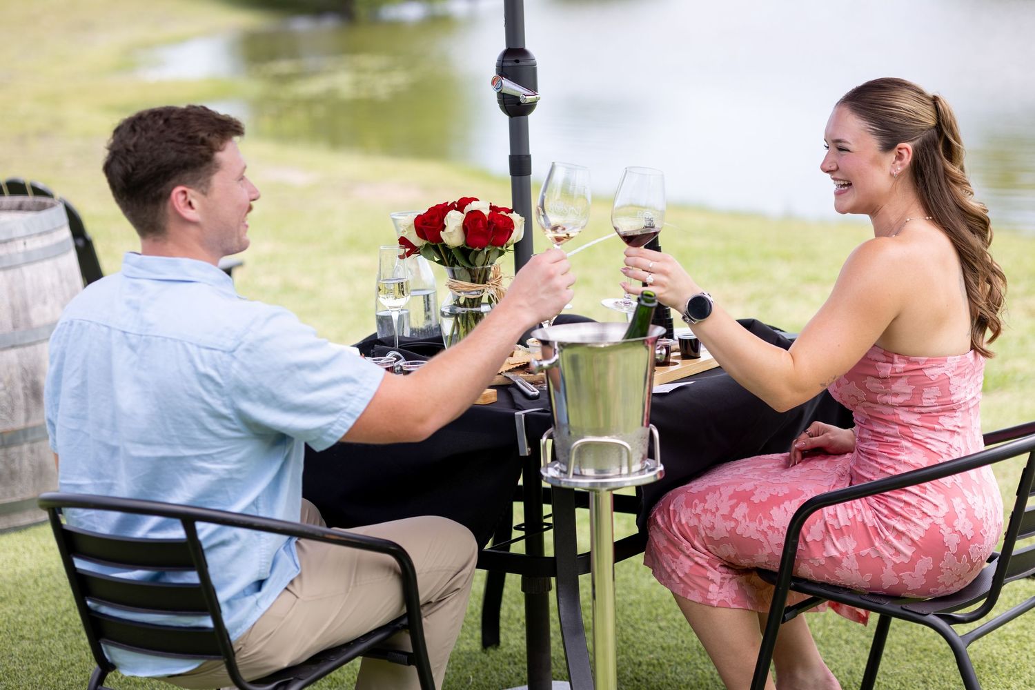 Couple toasting wine glasses at romantic outdoor lakeside dinner table with red roses.