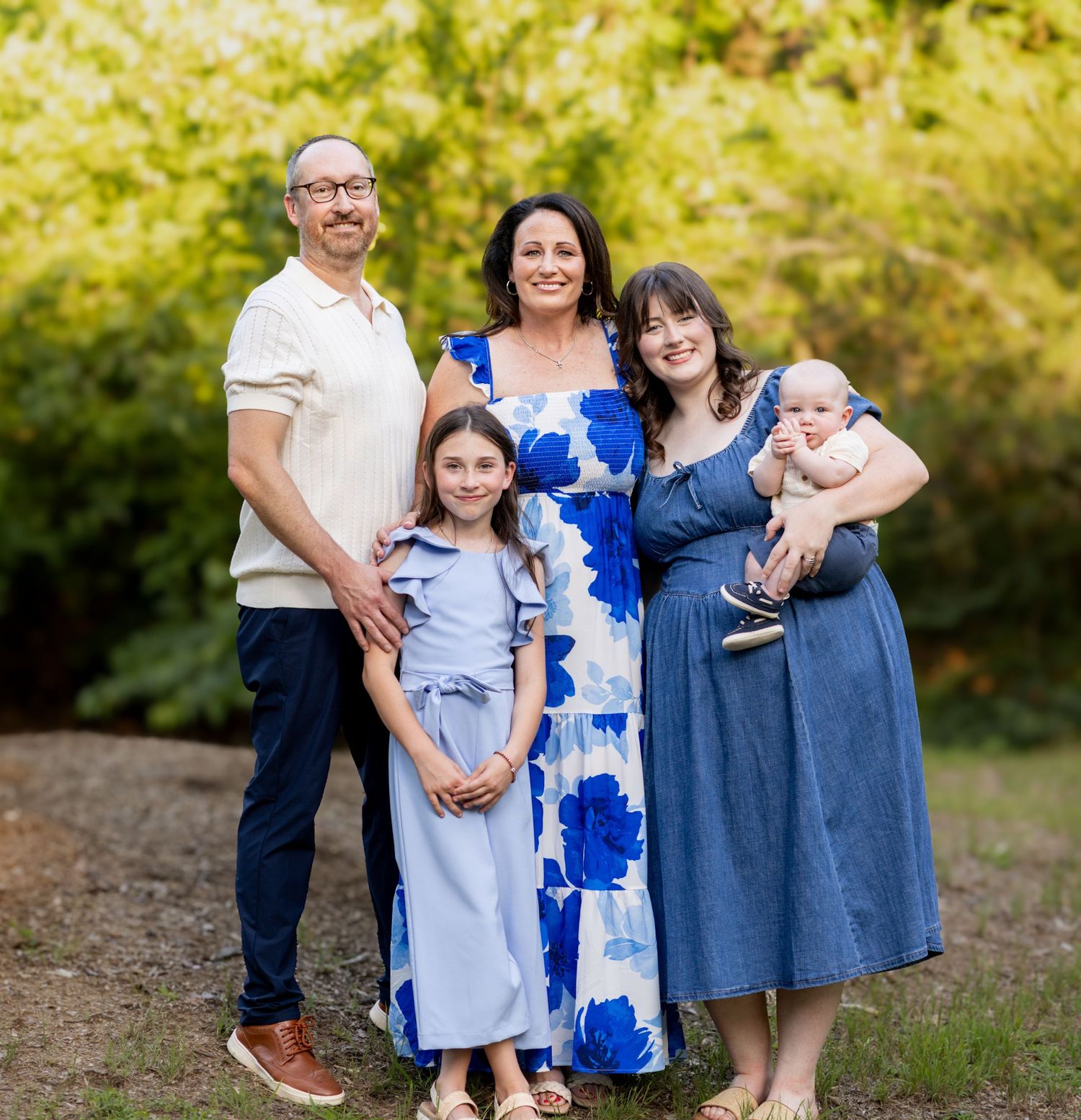 Family of five posing outdoors, smiling together among green trees in coordinating blue outfits.