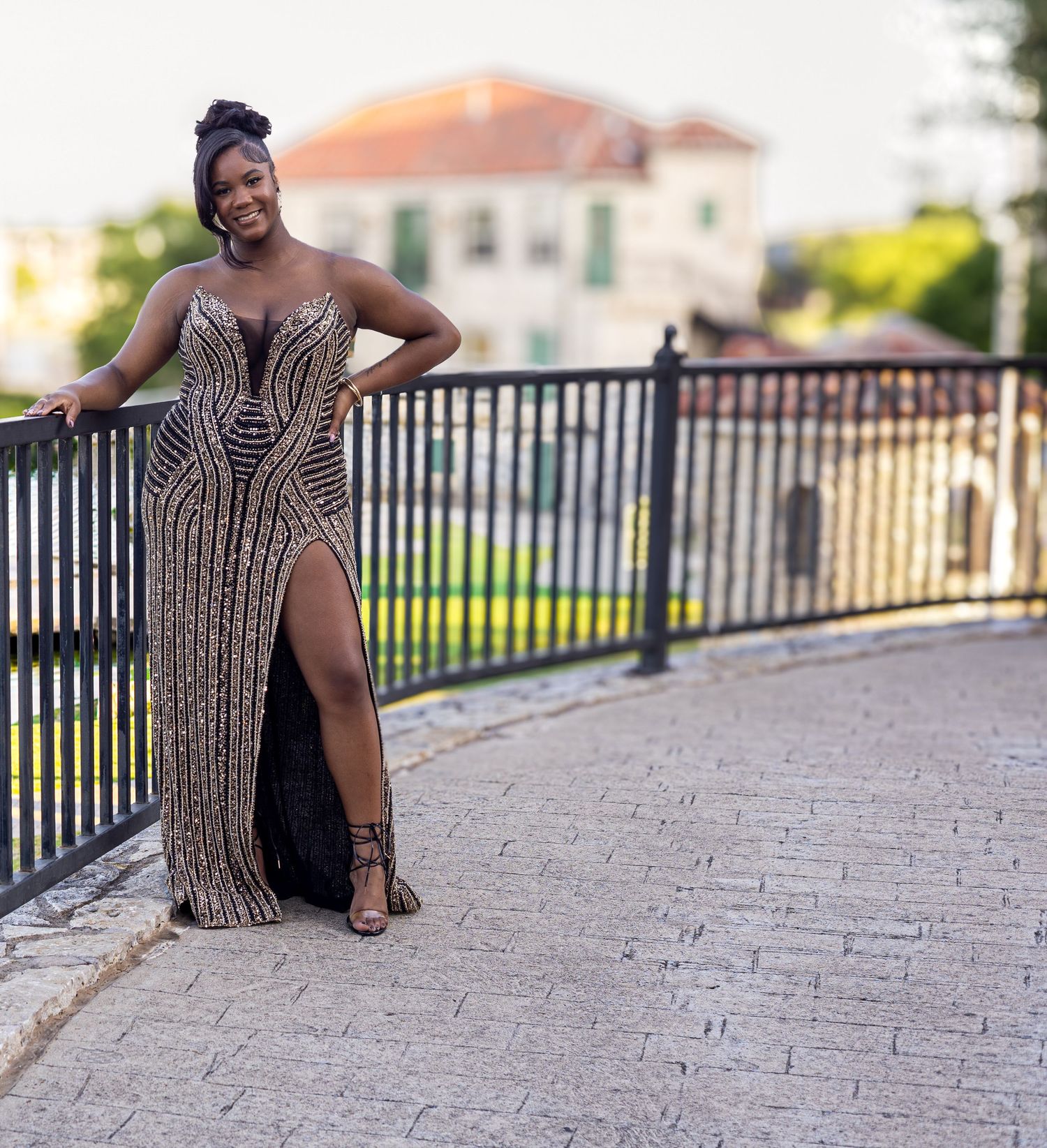 Woman in gold beaded slit gown poses confidently on brick walkway at sunset.
