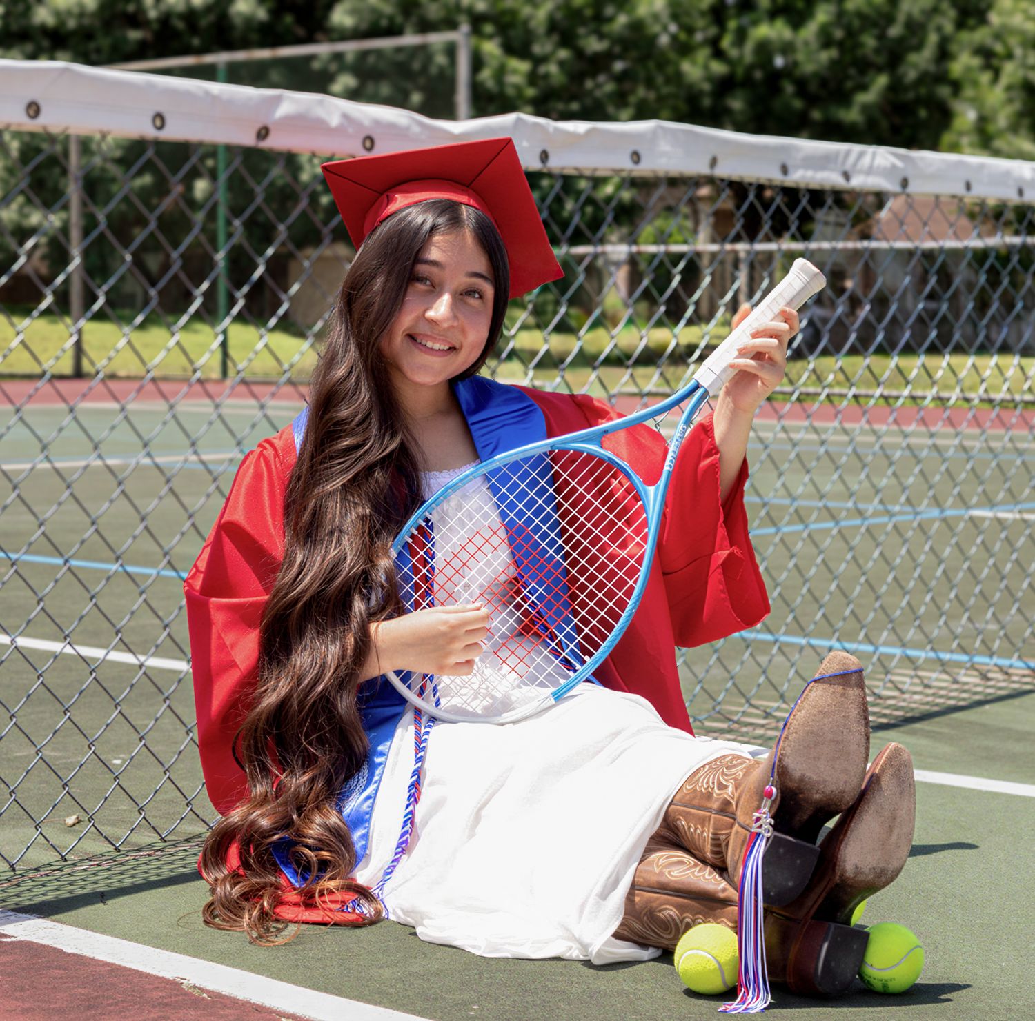 A young woman in red graduation cap and gown sits on a tennis court holding a diploma and blue tennis racket.