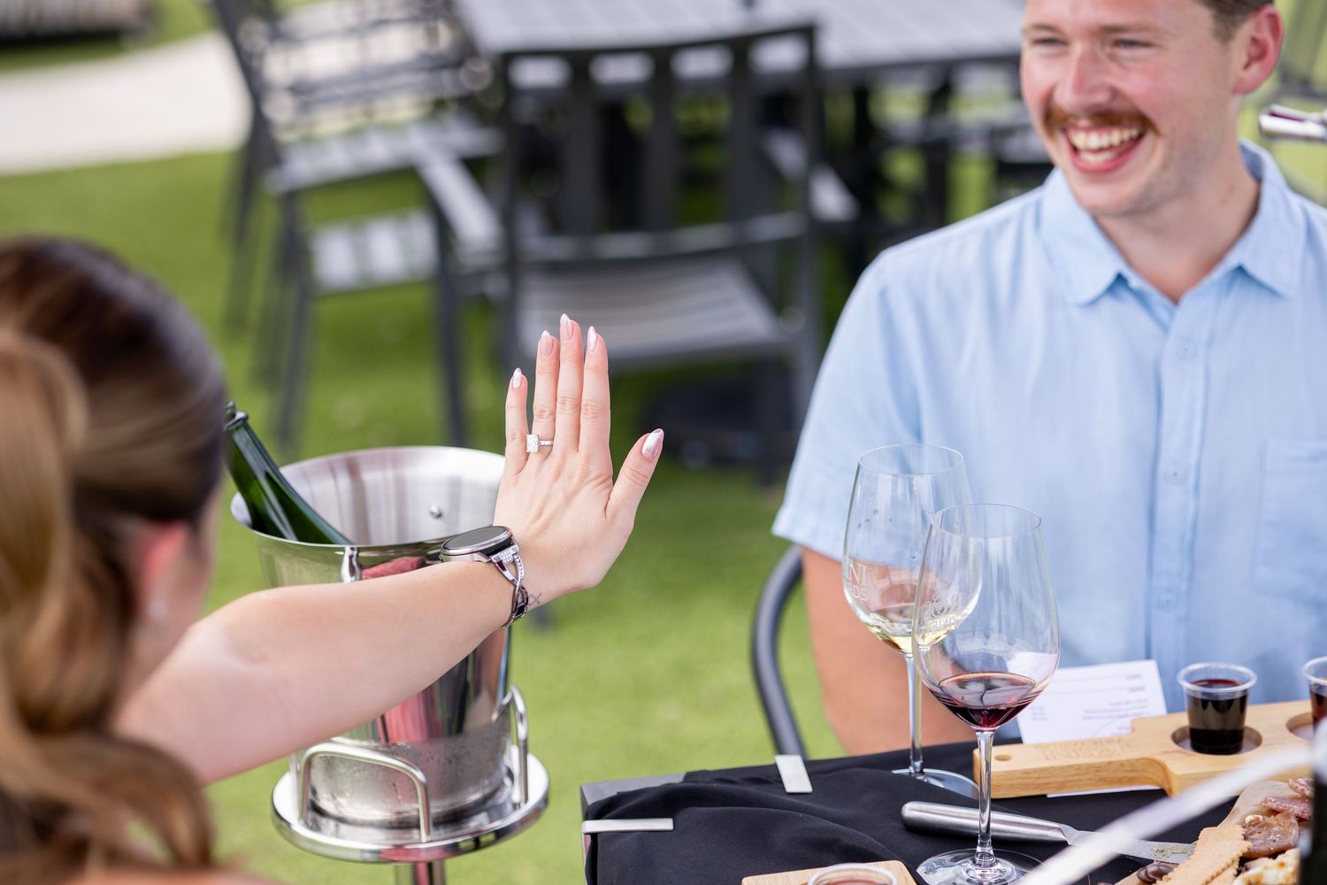 Two people enjoying wine tasting outdoors, sharing a high-five over a table with red wine glasses and a wine bucket.