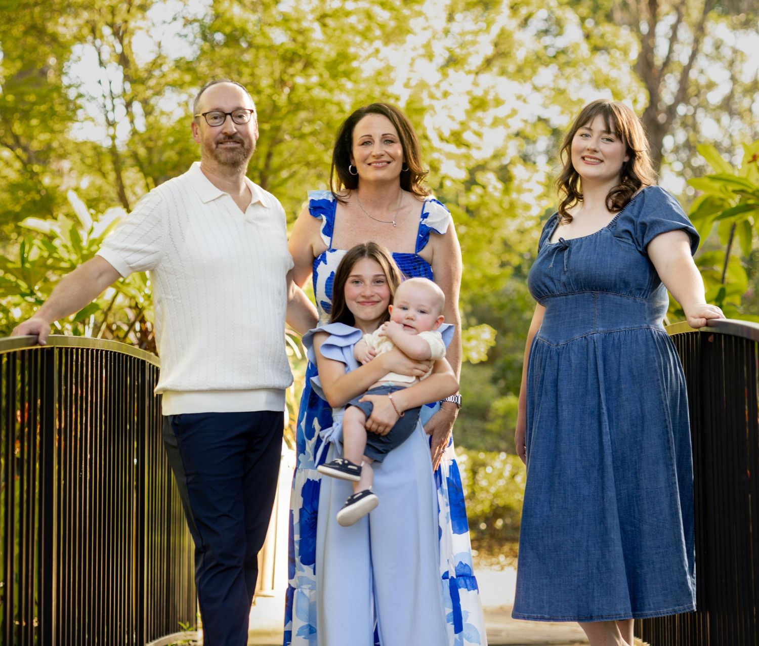 Family of five posing outdoors in coordinating blue and white outfits, with lush green trees in the background.