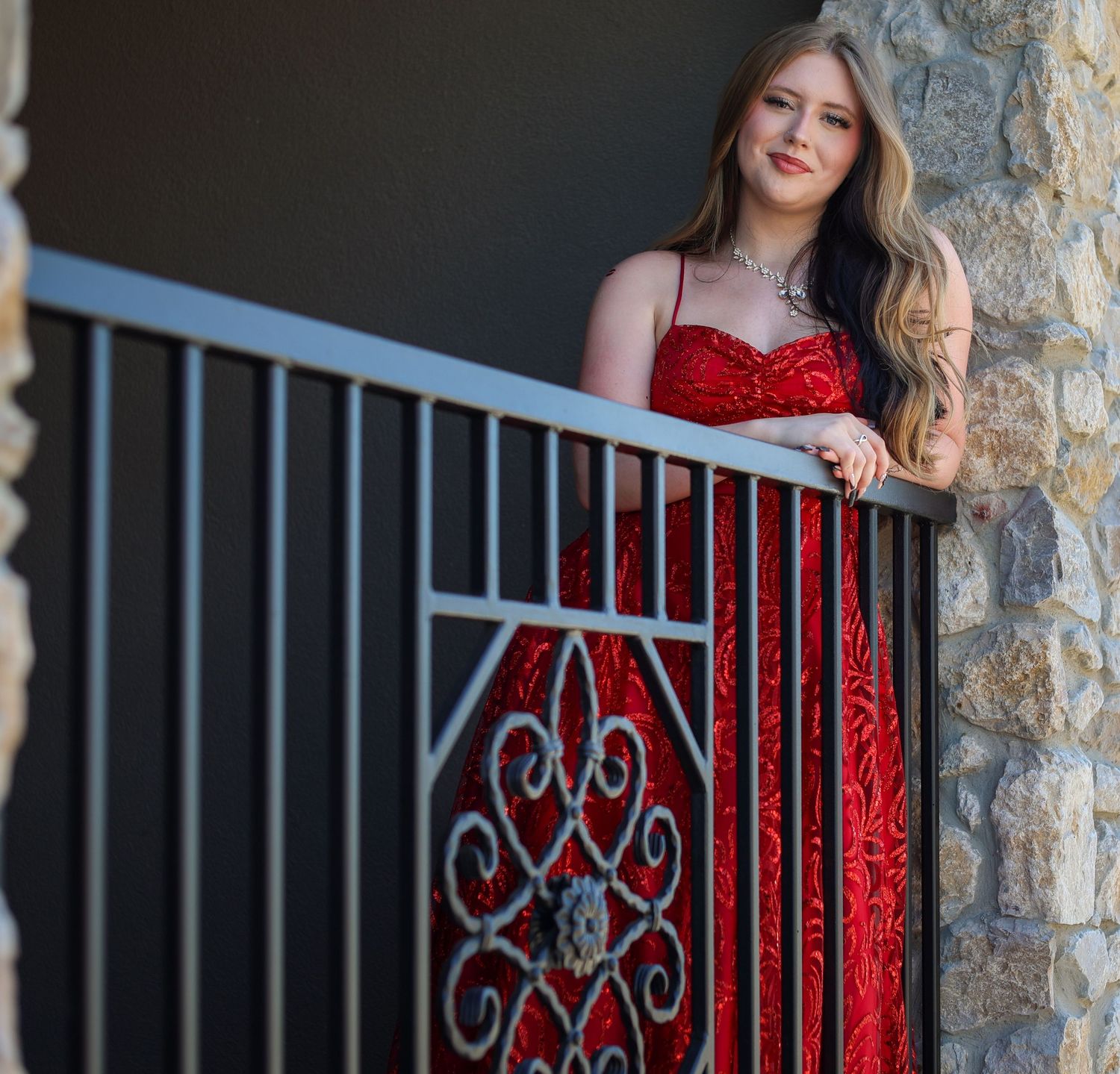 Young woman in red sequin gown leans on iron railing at stone building.