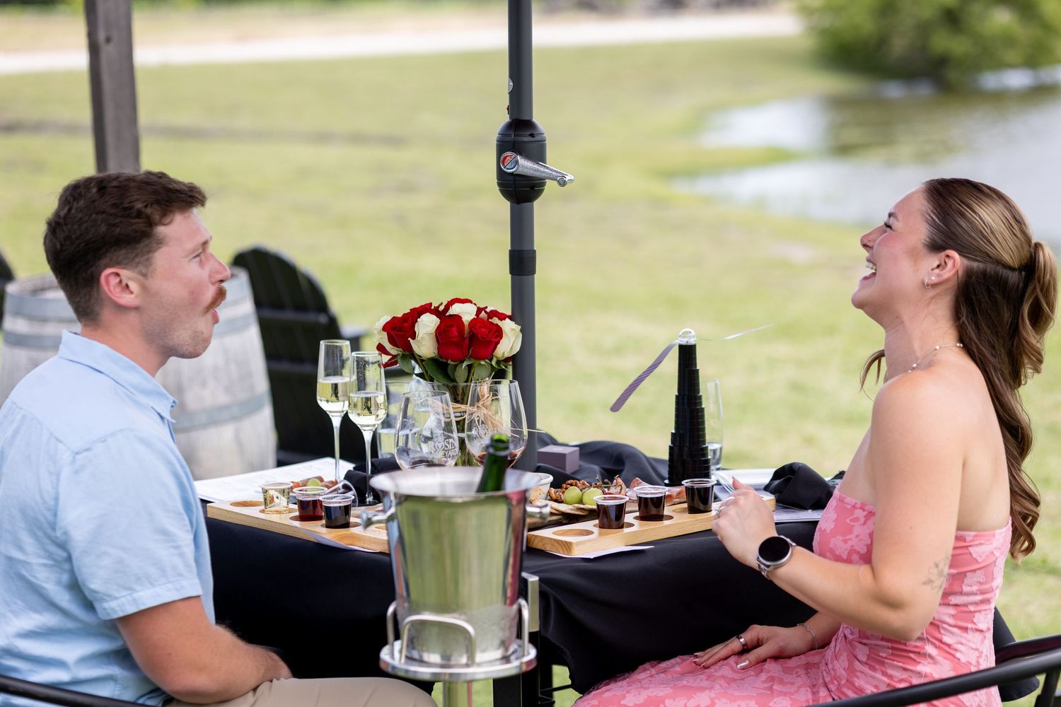 Couple enjoying a romantic outdoor dining experience with wine, roses, and gourmet food by a scenic waterfront.