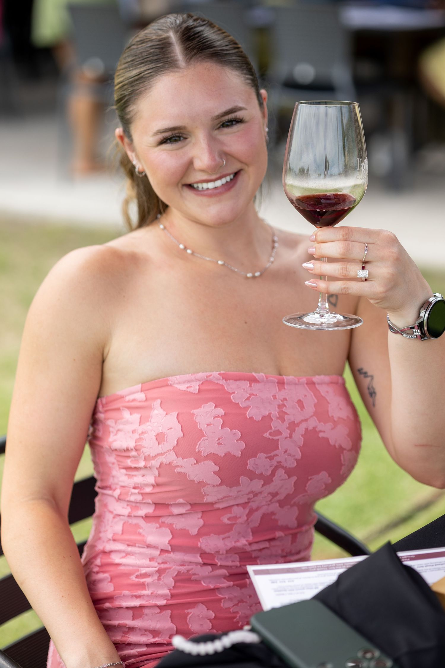 Smiling young woman in pink floral strapless dress raising a glass of red wine outdoors.